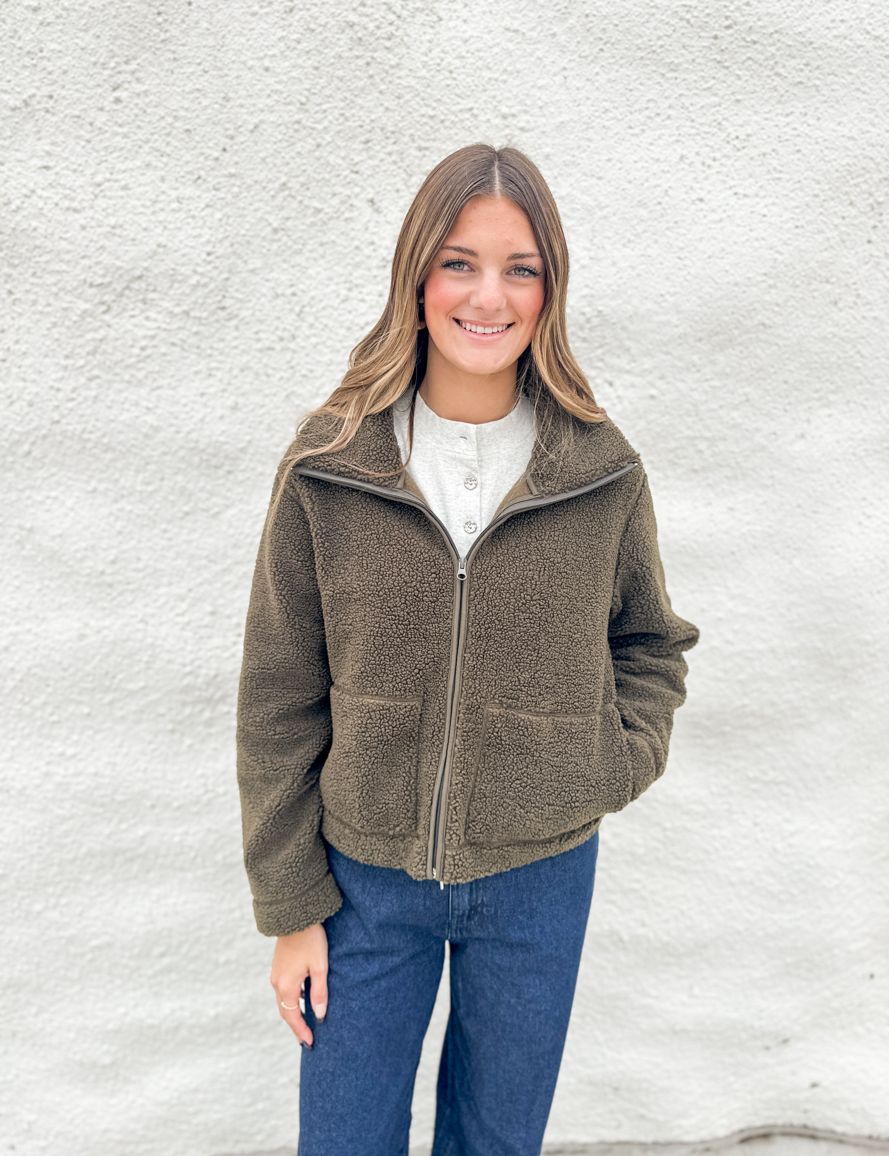A woman smiling while wearing an olive brown fleece zip-up jacket from Kut from the Kloth over a white henley shirt and blue jeans, standing against a white textured wall.