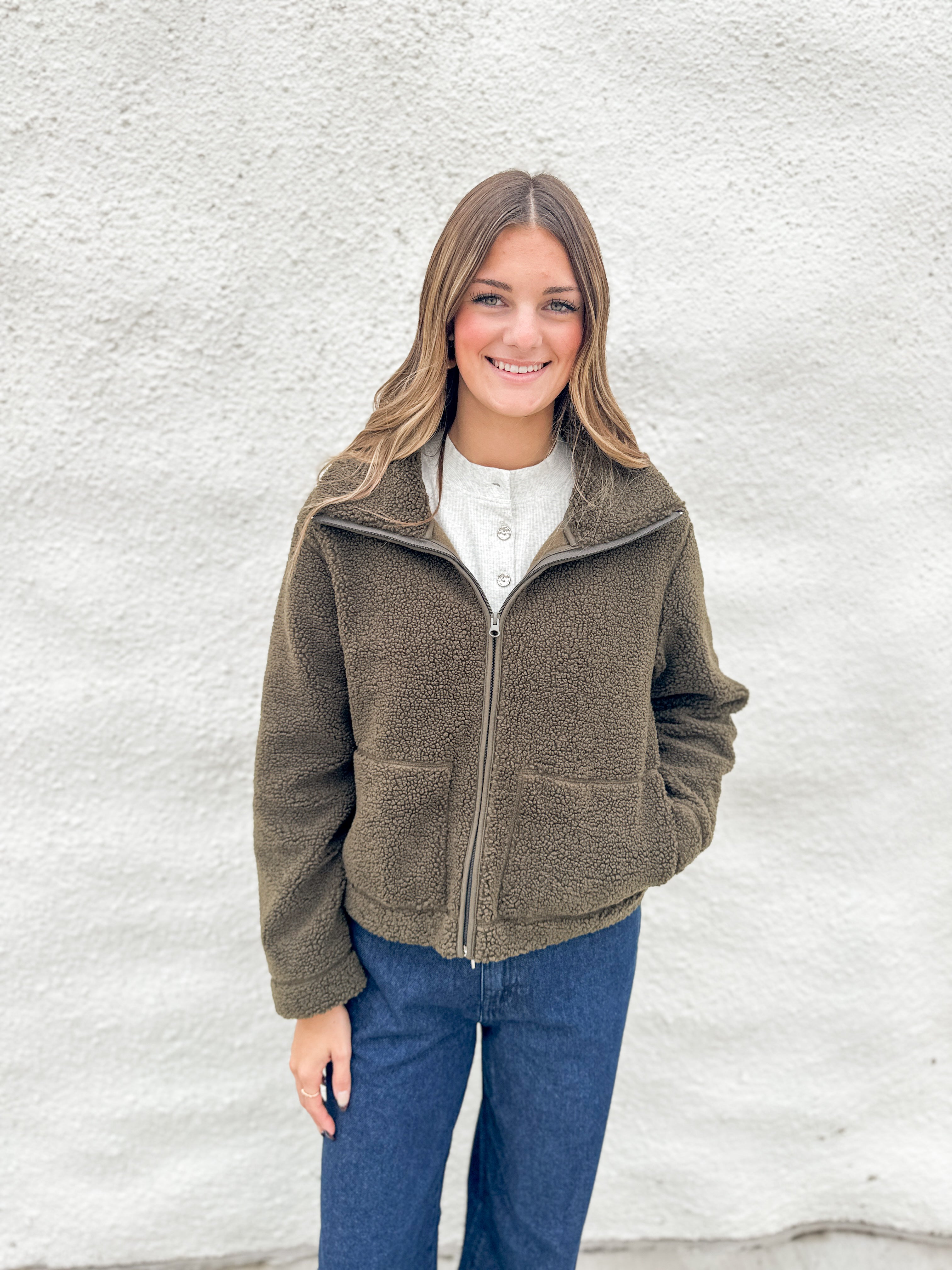 A woman smiling while wearing an olive brown fleece zip-up jacket from Kut from the Kloth over a white henley shirt and blue jeans, standing against a white textured wall.