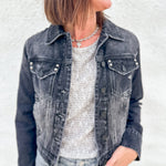 A woman wearing a washed black denim jacket with stud details over a textured light gray top and light blue jeans, standing against a white stucco wall.