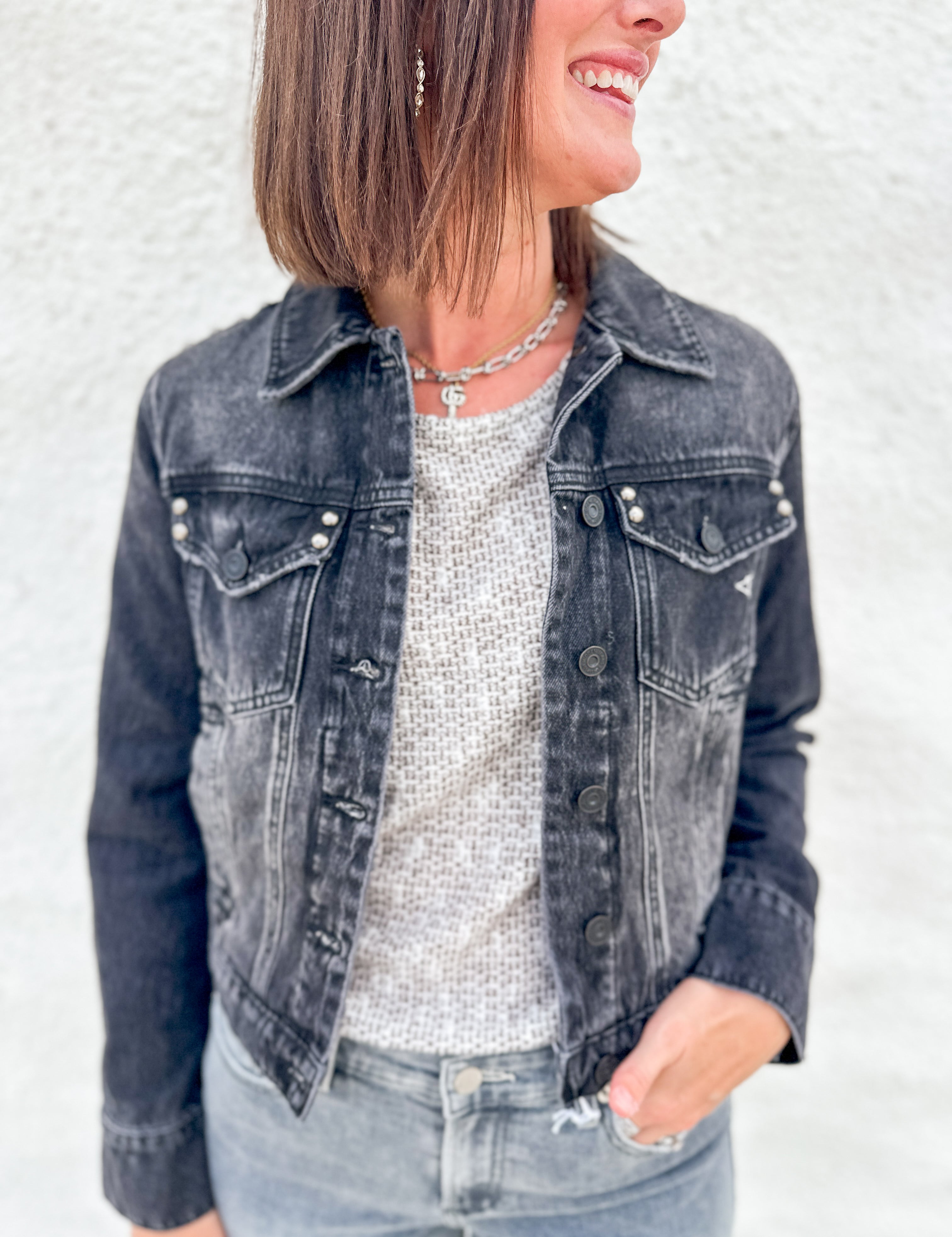 A woman wearing a washed black denim jacket with stud details over a textured light gray top and light blue jeans, standing against a white stucco wall.