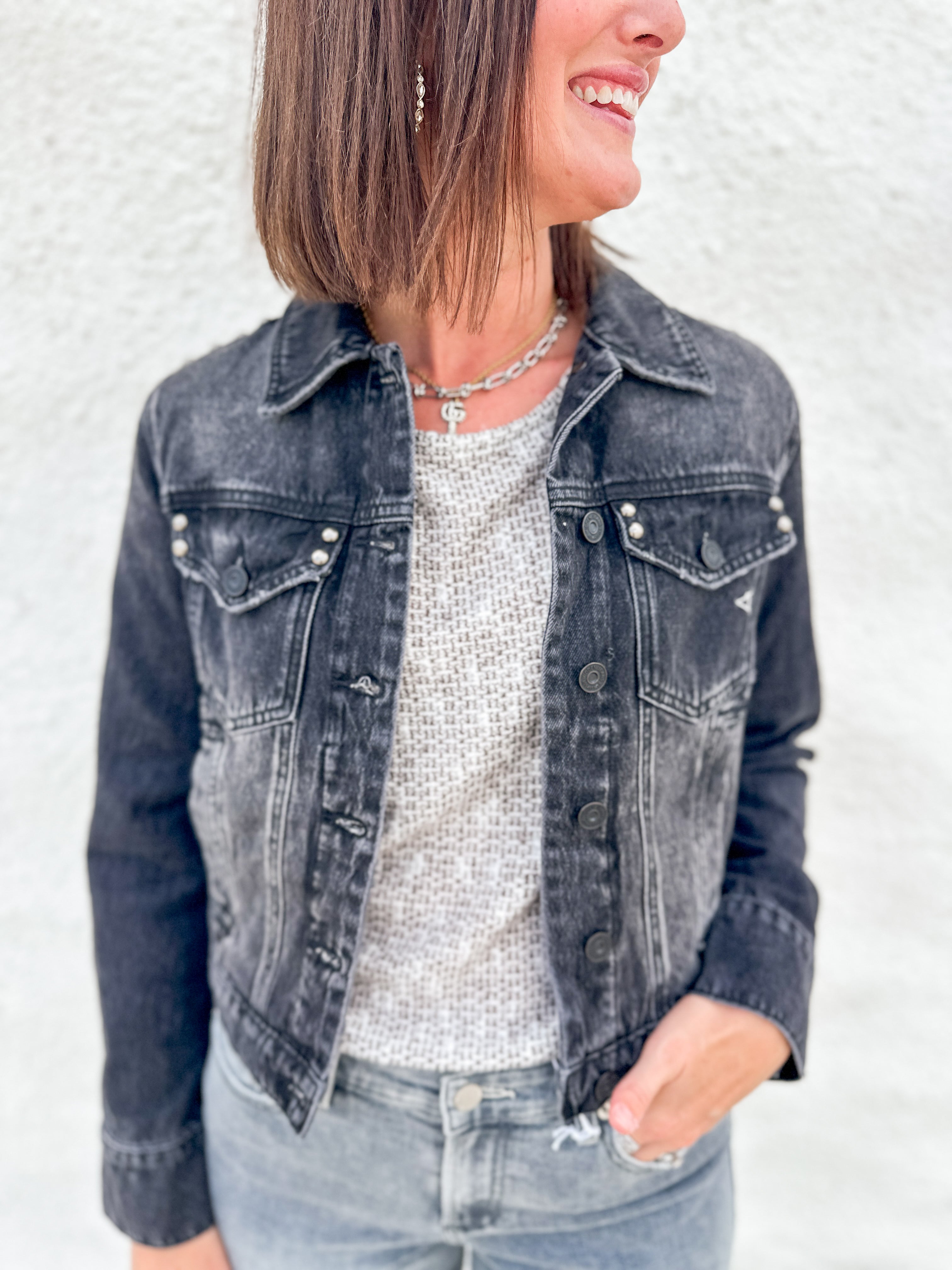 A woman wearing a washed black denim jacket with stud details over a textured light gray top and light blue jeans, standing against a white stucco wall.