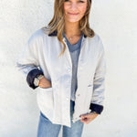A woman wearing a light beige utility-style jacket with dark navy cuffs and collar, layered over a blue-gray striped sweater and blue jeans, standing against a white textured wall.