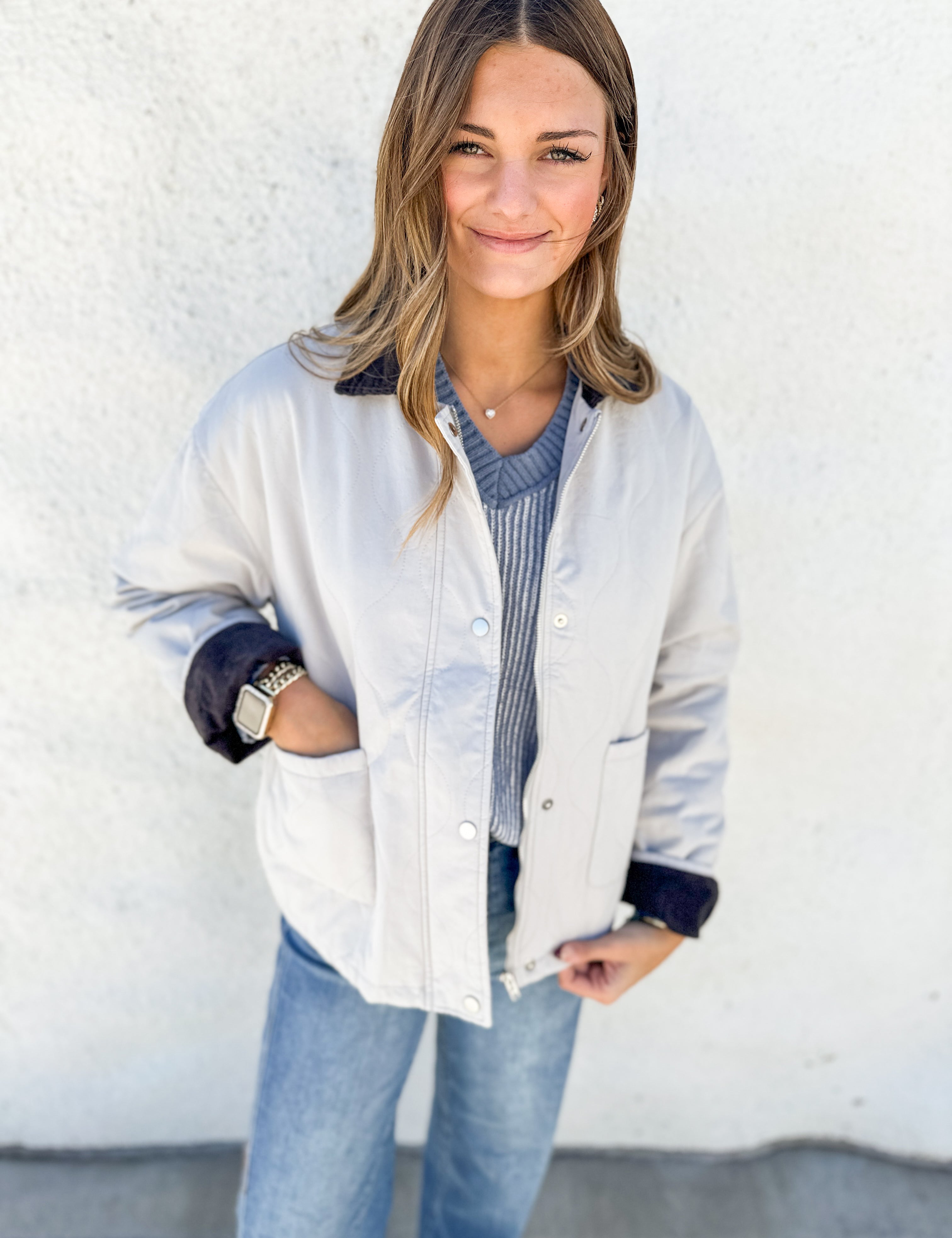 A woman wearing a light beige utility-style jacket with dark navy cuffs and collar, layered over a blue-gray striped sweater and blue jeans, standing against a white textured wall.