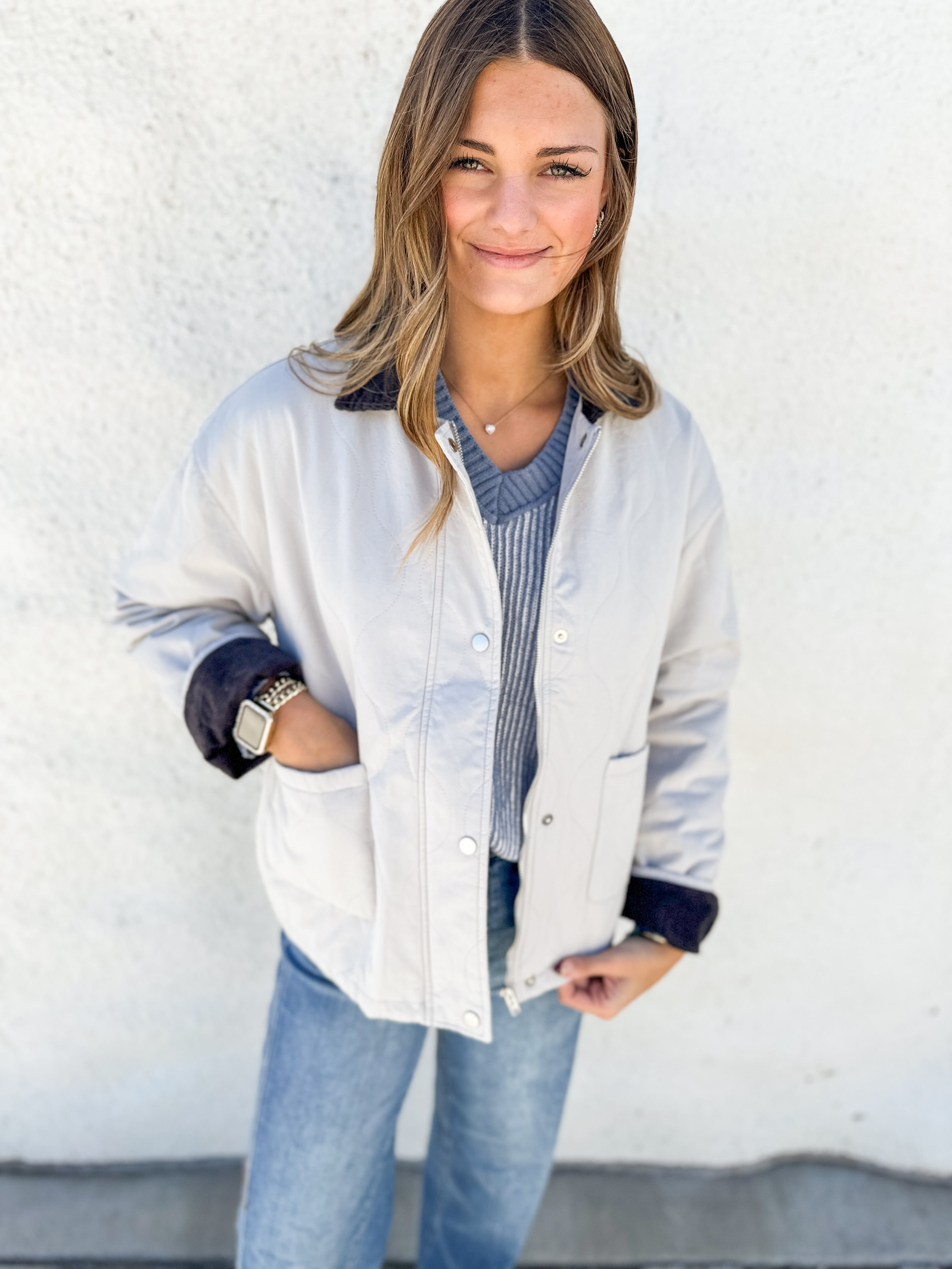 A woman wearing a light beige utility-style jacket with dark navy cuffs and collar, layered over a blue-gray striped sweater and blue jeans, standing against a white textured wall.