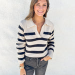A woman wearing a cream and black horizontally striped knit polo sweater, paired with dark gray jeans, standing against a white textured wall.