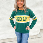 A girl wearing a green and gold sweater that says Green Bay with white stripes on the elbows wearing blue jeans against a white wall.