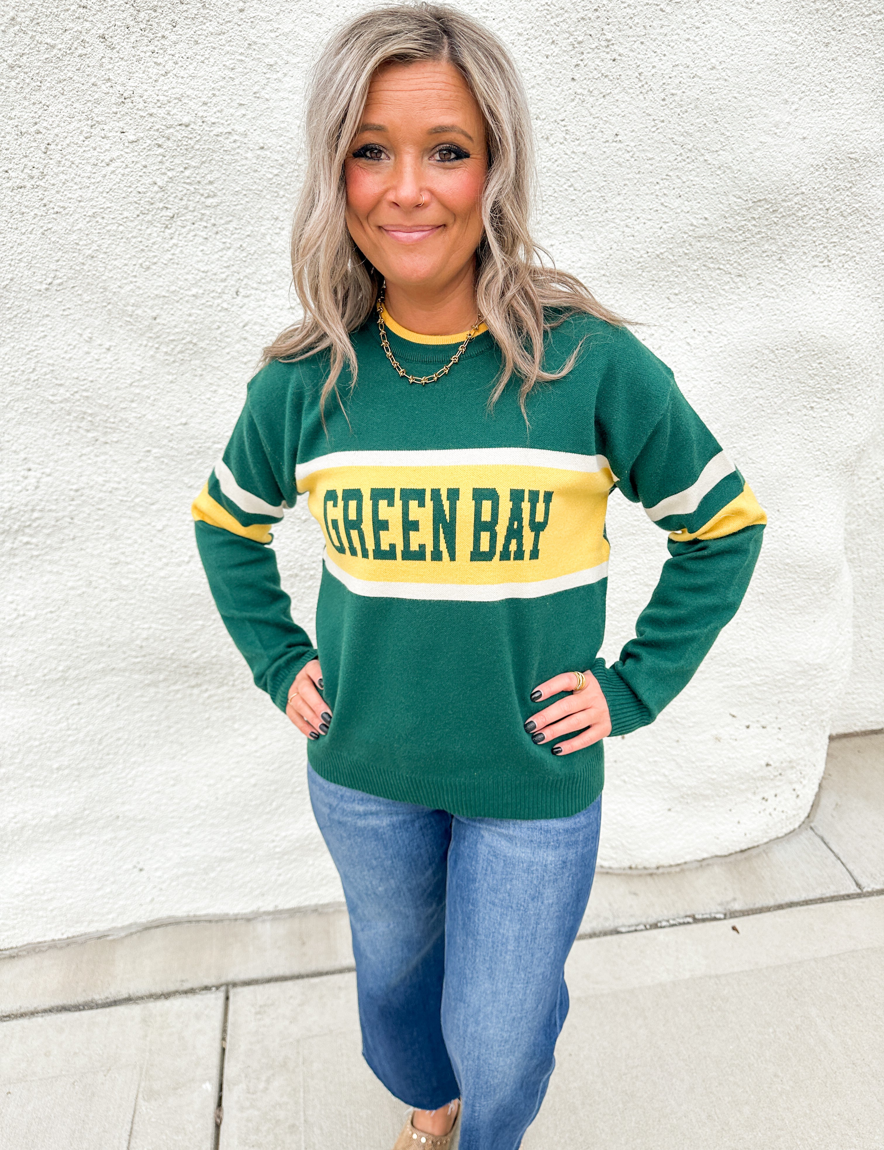 A girl wearing a green and gold sweater that says Green Bay with white stripes on the elbows wearing blue jeans against a white wall.