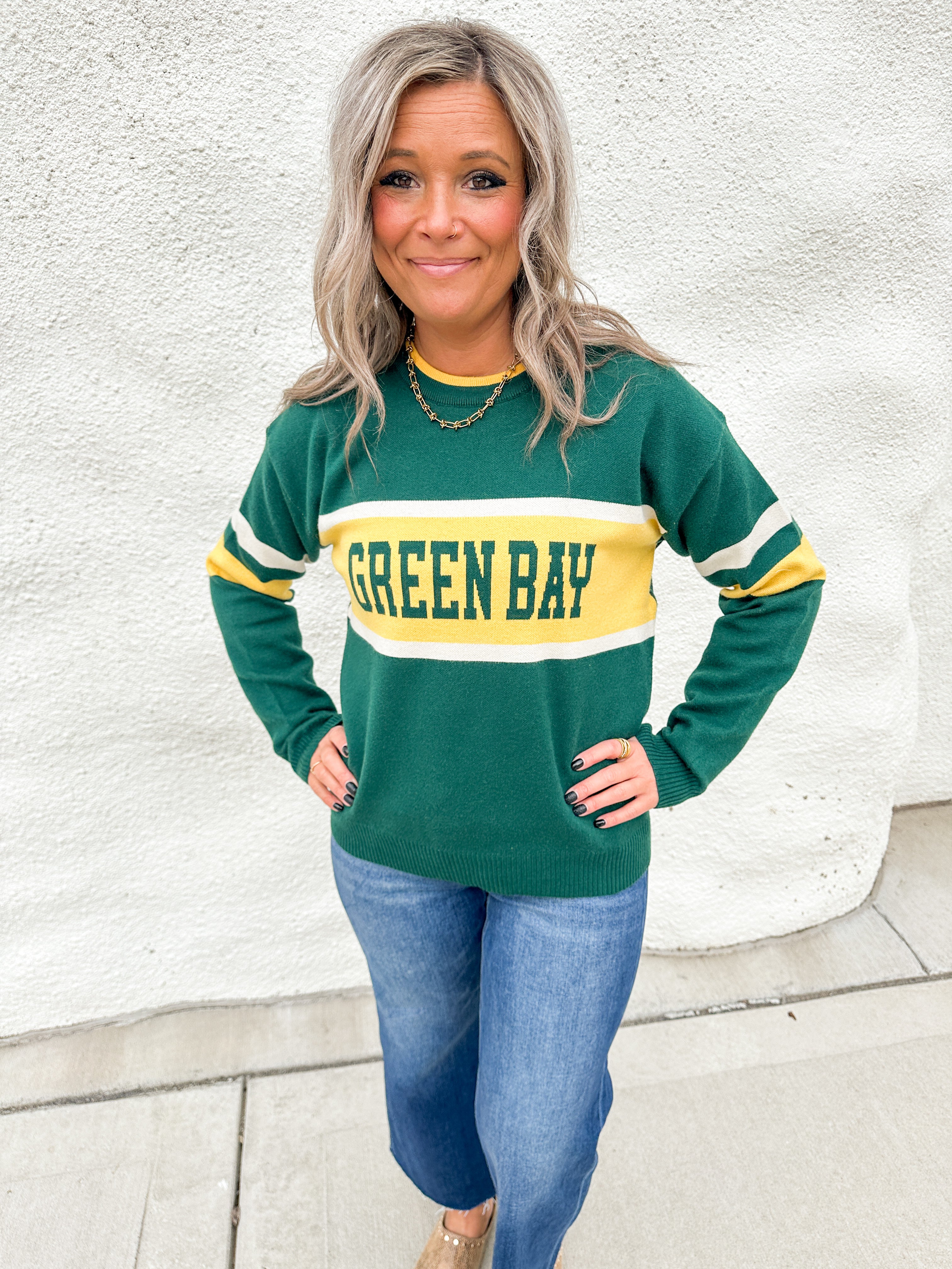 A girl wearing a green and gold sweater that says Green Bay with white stripes on the elbows wearing blue jeans against a white wall.