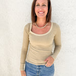 A smiling woman with brown hair stands against a white textured wall, wearing a beige long-sleeve scoop-neck ribbed top and blue jeans.