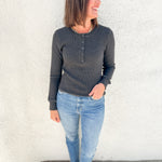 A woman wearing a dark charcoal gray long-sleeve henley shirt, light blue jeans, and brown flat shoes, standing against a white stucco wall.