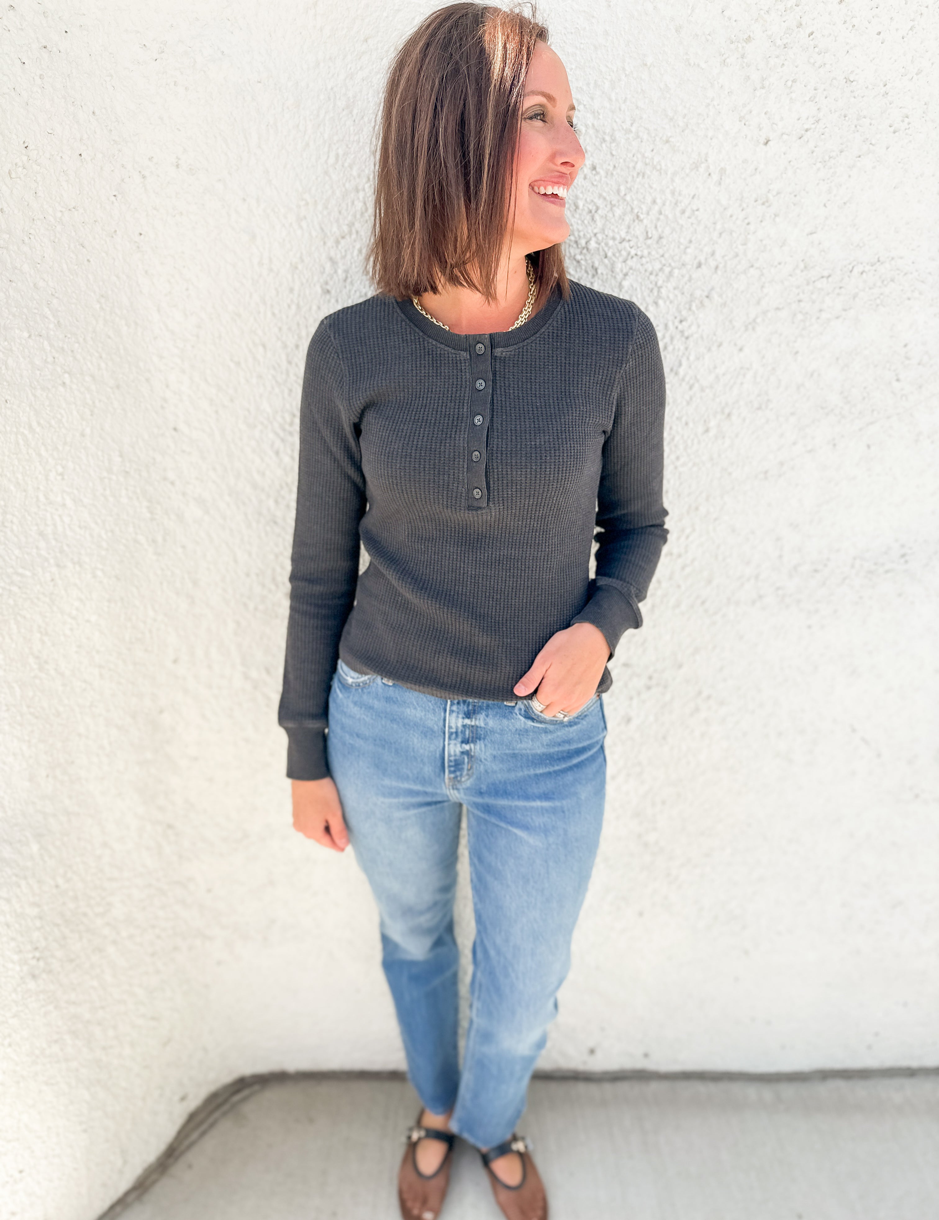 A woman wearing a dark charcoal gray long-sleeve henley shirt, light blue jeans, and brown flat shoes, standing against a white stucco wall.