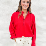 A woman smiling while wearing a bright red long-sleeve quarter-zip pullover and cream barrel drawstring pants with brown polka dots, standing against a white textured wall.