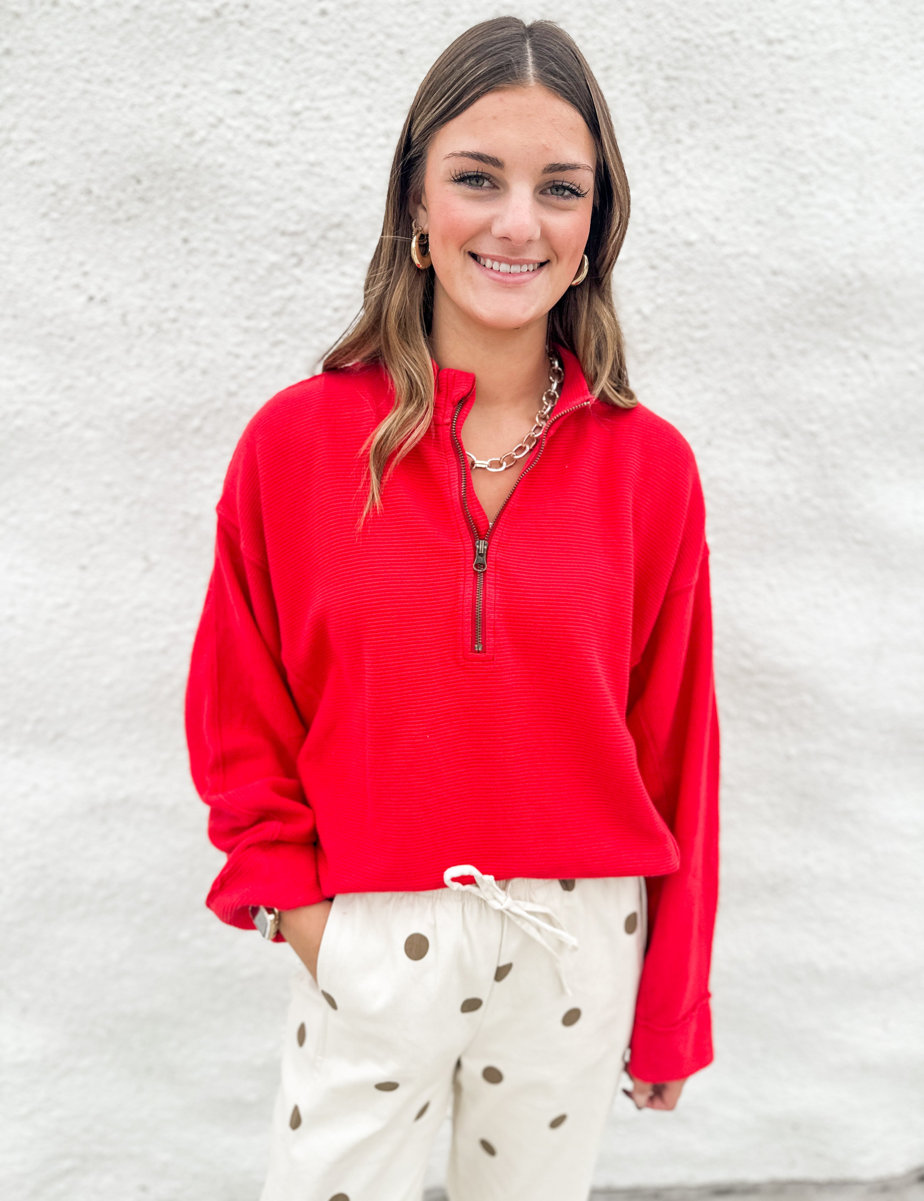 A woman smiling while wearing a bright red long-sleeve quarter-zip pullover and cream barrel drawstring pants with brown polka dots, standing against a white textured wall.