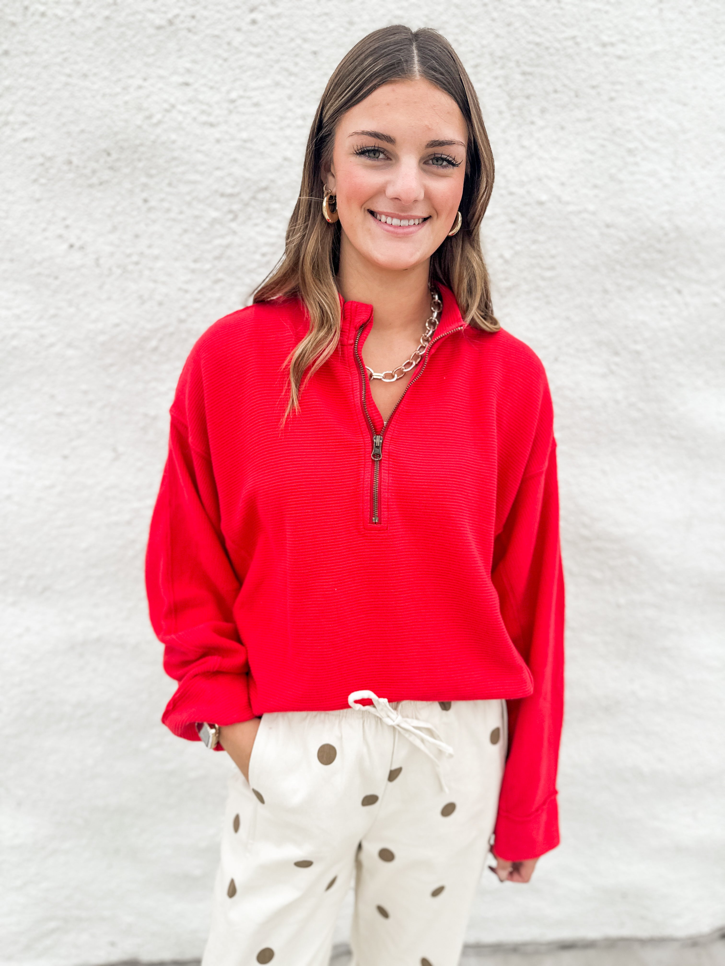 A woman smiling while wearing a bright red long-sleeve quarter-zip pullover and cream barrel drawstring pants with brown polka dots, standing against a white textured wall.