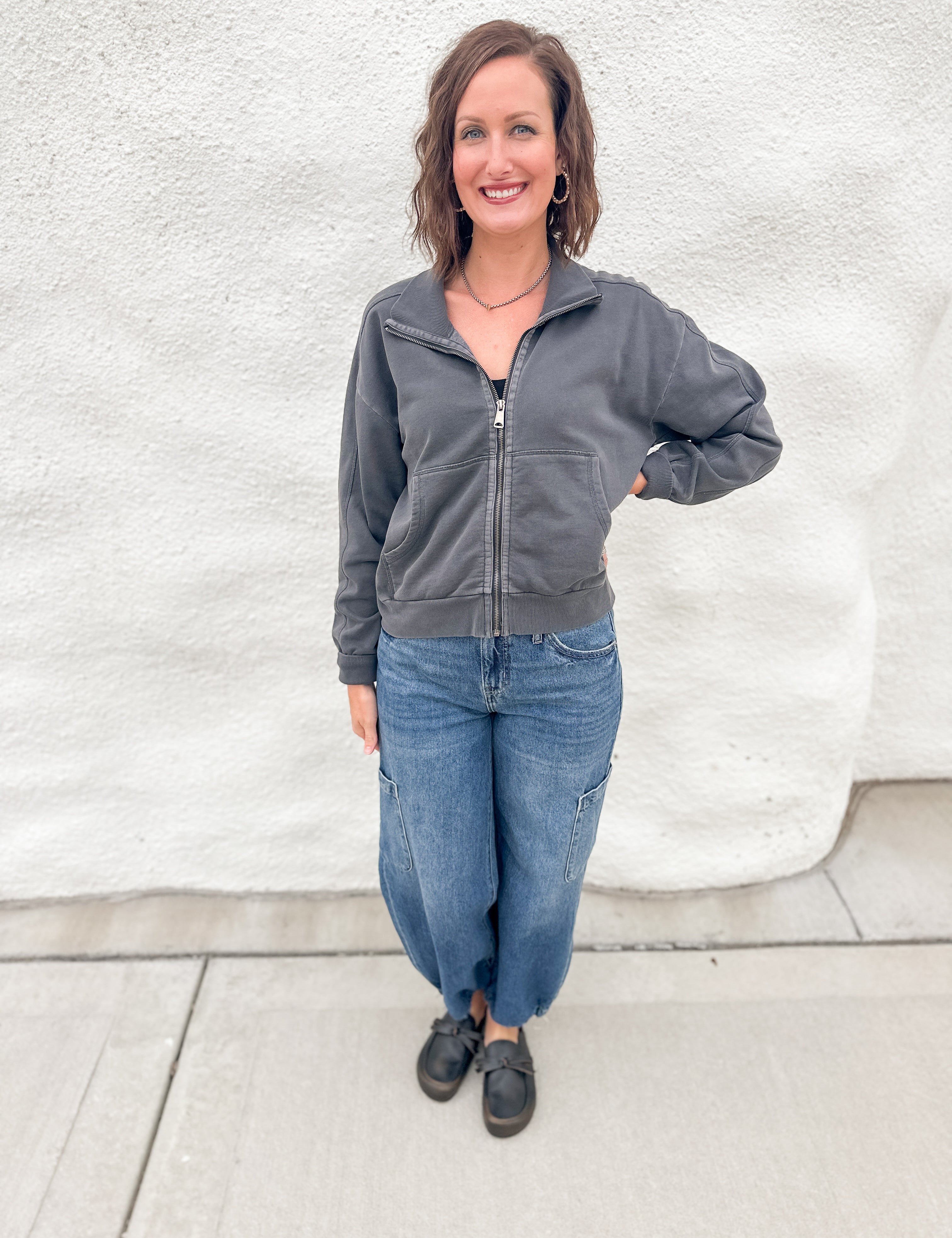 A woman wearing a gray zippered sweatshirt and blue denim joggers with distressed black leather mule clogs standing in front of a white wall.