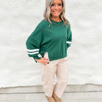 A girl wearing a green sweatshirt with white stripes on sleeves and notches around bottom hem wearing fawn print barrel leg jeans standing in front of a white wall.