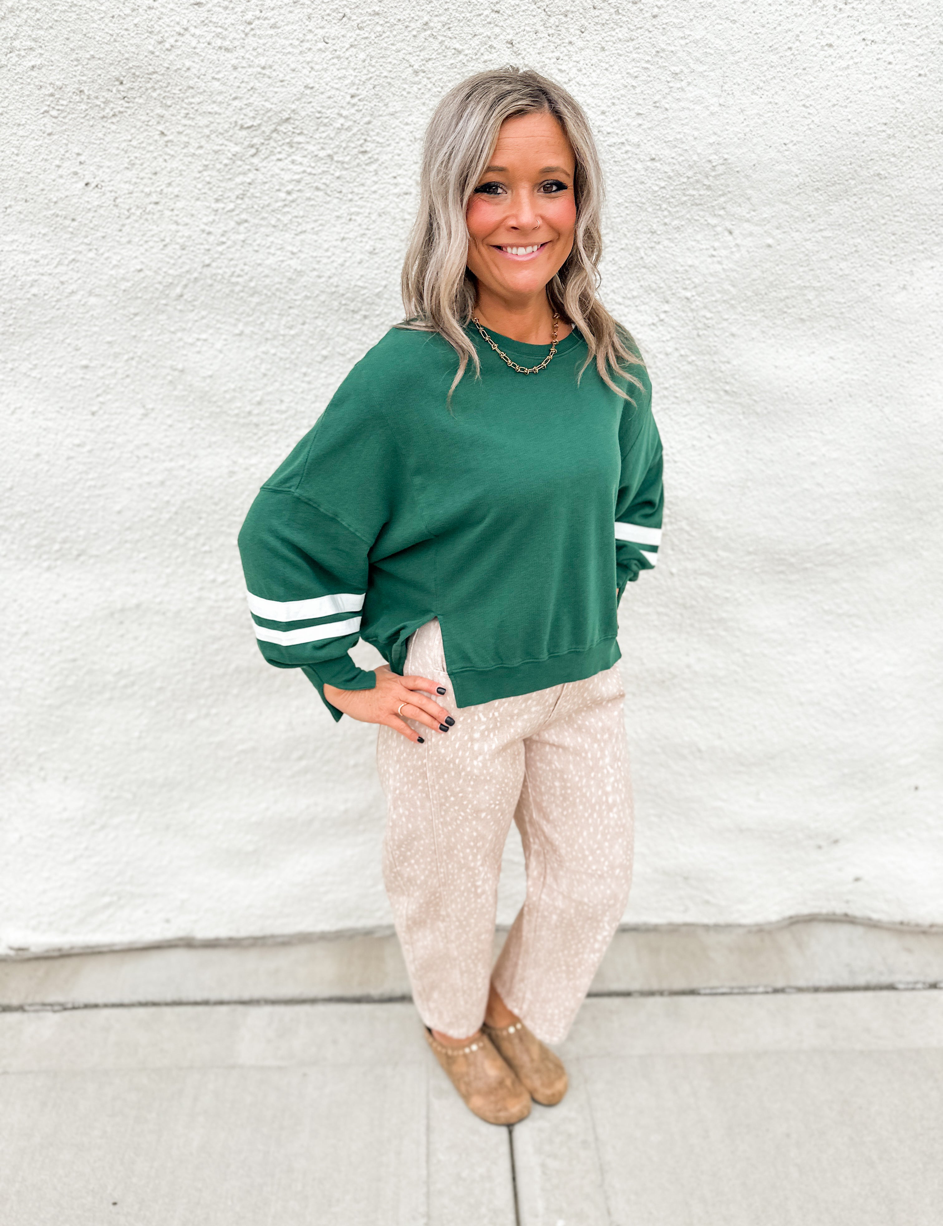 A girl wearing a green sweatshirt with white stripes on sleeves and notches around bottom hem wearing fawn print barrel leg jeans standing in front of a white wall.