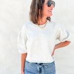 A woman wearing a light heather gray short-sleeve crewneck sweatshirt and blue jeans, standing against a textured white wall.