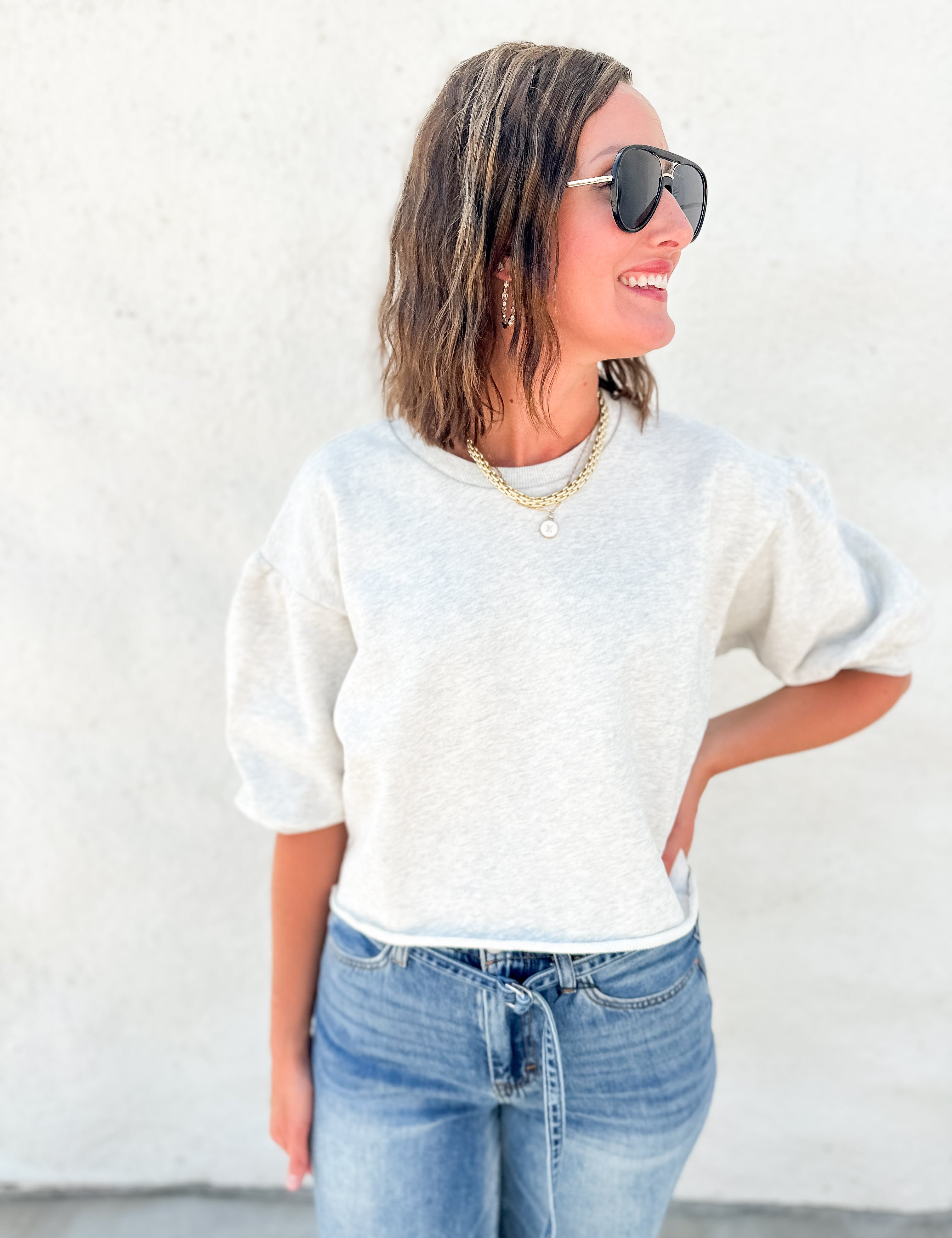 A woman wearing a light heather gray short-sleeve crewneck sweatshirt and blue jeans, standing against a textured white wall.