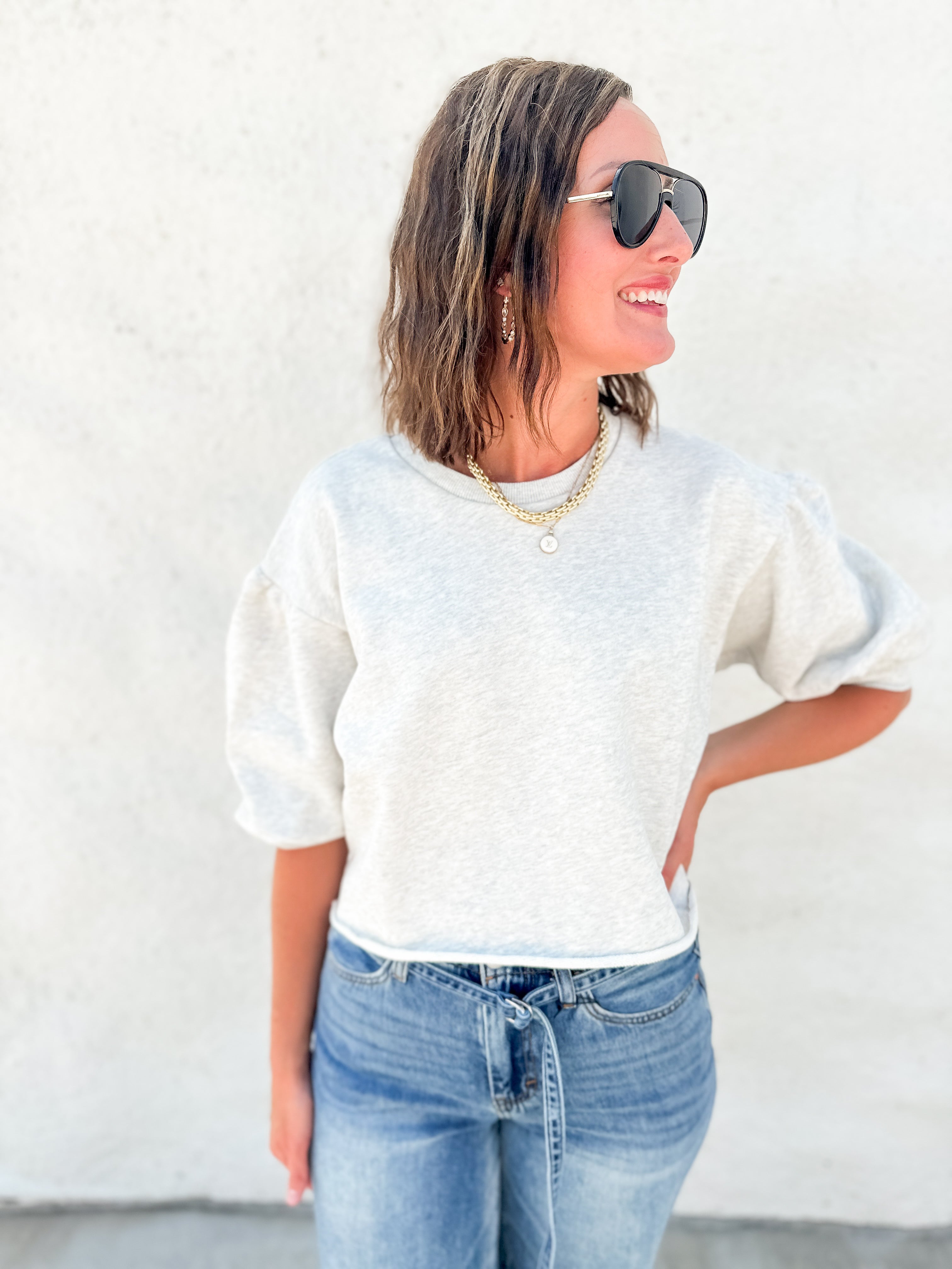 A woman wearing a light heather gray short-sleeve crewneck sweatshirt and blue jeans, standing against a textured white wall.