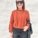 A woman wearing a rust-orange cable-knit mock-neck sweater, dark gray jeans, black sunglasses, and a gold chain necklace, holding a black bag against a textured white wall.
