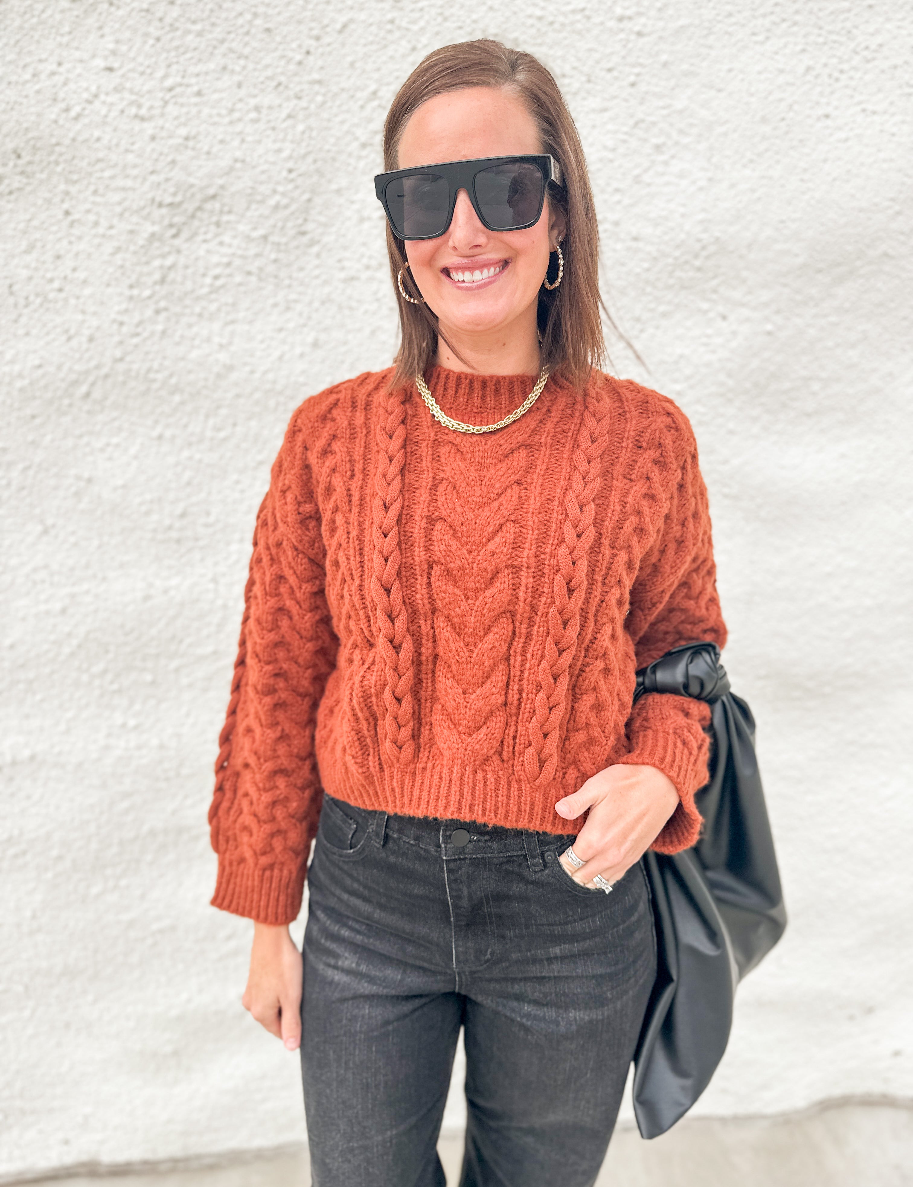A woman wearing a rust-orange cable-knit mock-neck sweater, dark gray jeans, black sunglasses, and a gold chain necklace, holding a black bag against a textured white wall.