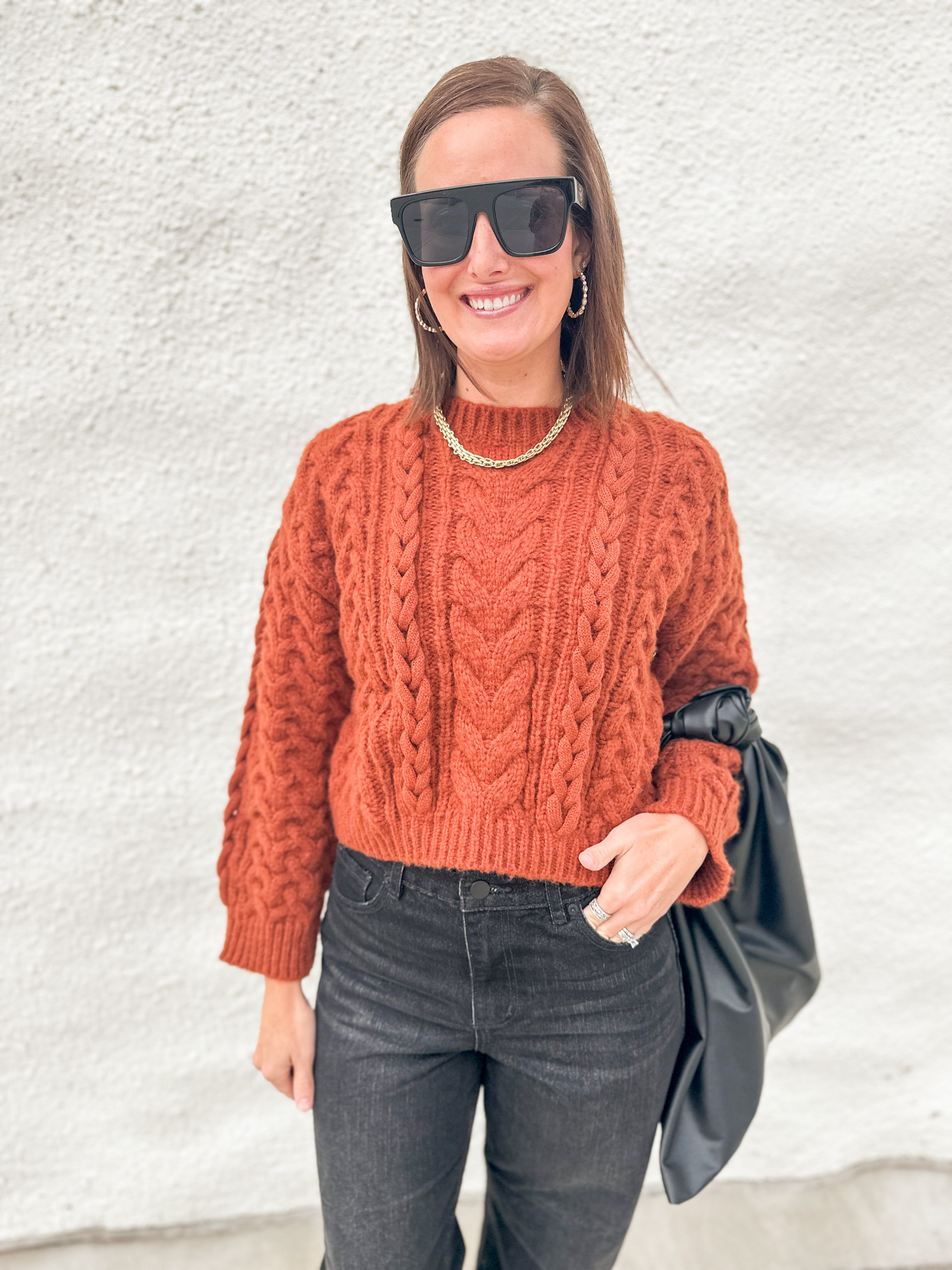 A woman wearing a rust-orange cable-knit mock-neck sweater, dark gray jeans, black sunglasses, and a gold chain necklace, holding a black bag against a textured white wall.