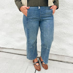 A person wearing light blue cropped denim jeans with front patch pockets, pull on elastic waist and inner drawstring by Dear John styled with a dark olive green ribbed sweater, a beige jacket, and brown Mary Jane flats, standing against a white textured wall.