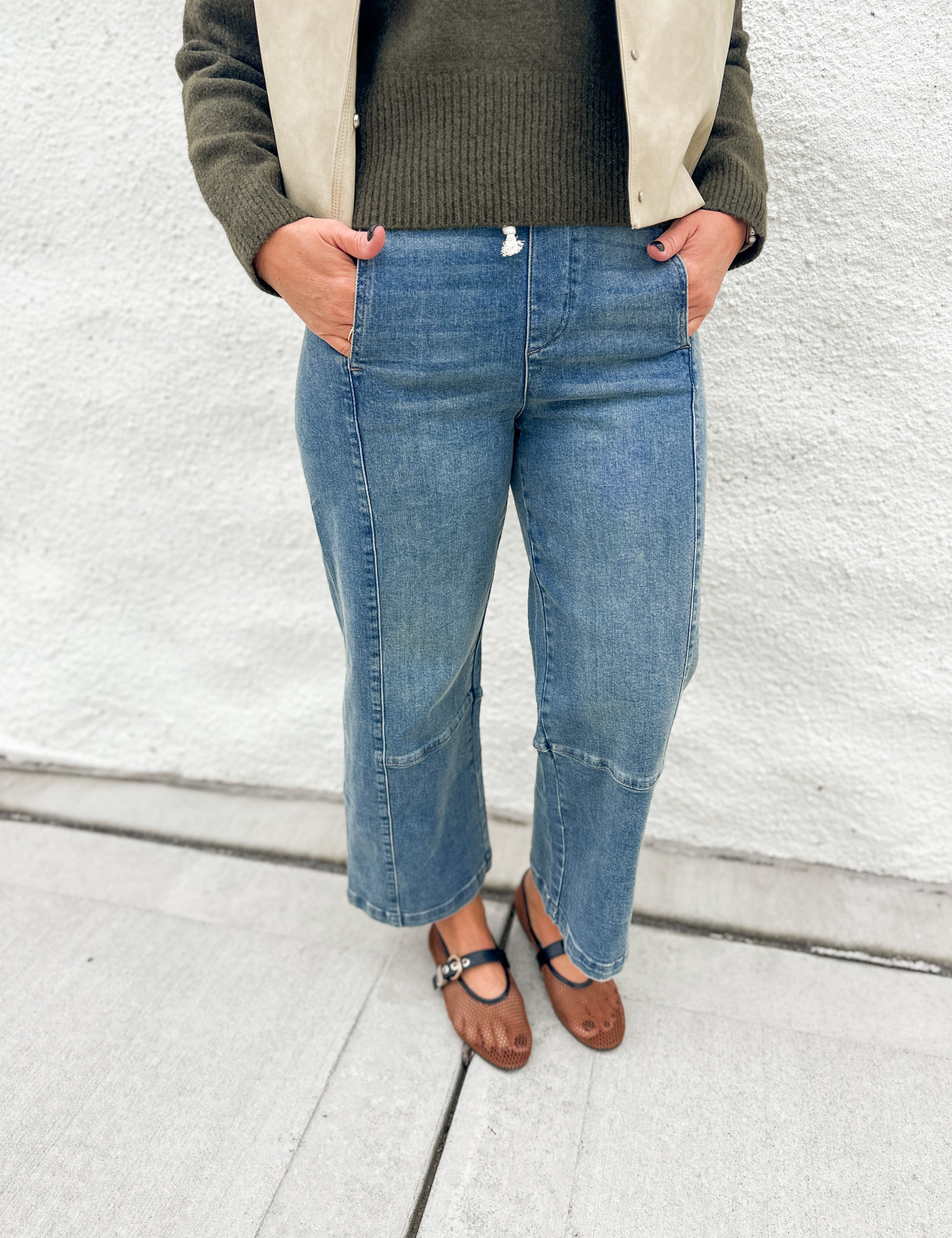 A person wearing light blue cropped denim jeans with front patch pockets, pull on elastic waist and inner drawstring by Dear John styled with a dark olive green ribbed sweater, a beige jacket, and brown Mary Jane flats, standing against a white textured wall.
