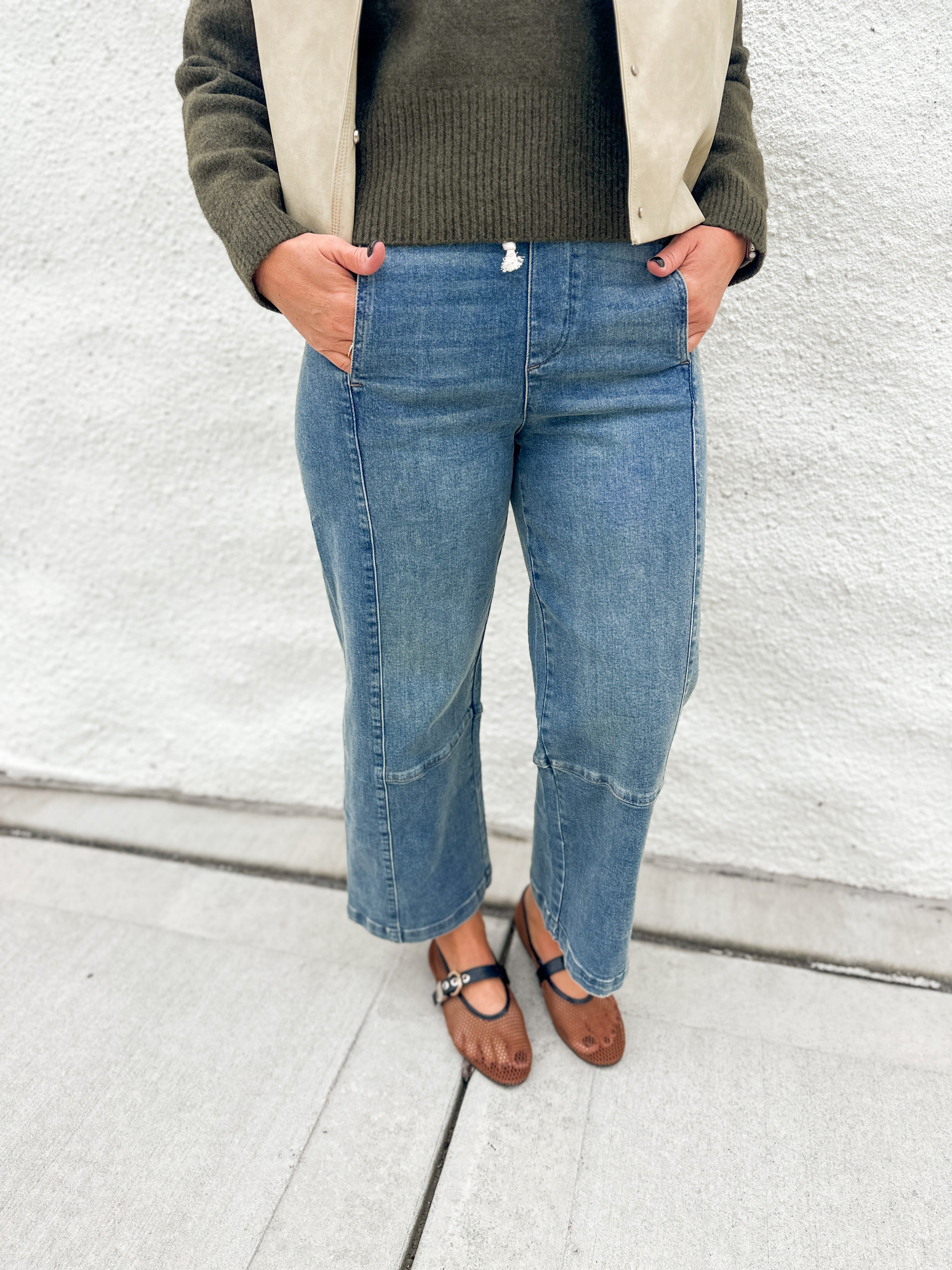 A person wearing light blue cropped denim jeans with front patch pockets, pull on elastic waist and inner drawstring by Dear John styled with a dark olive green ribbed sweater, a beige jacket, and brown Mary Jane flats, standing against a white textured wall.