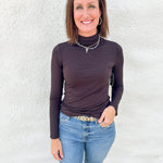 A woman wearing a dark brown long-sleeve mock neck turtleneck top and blue jeans, standing against a white textured wall.