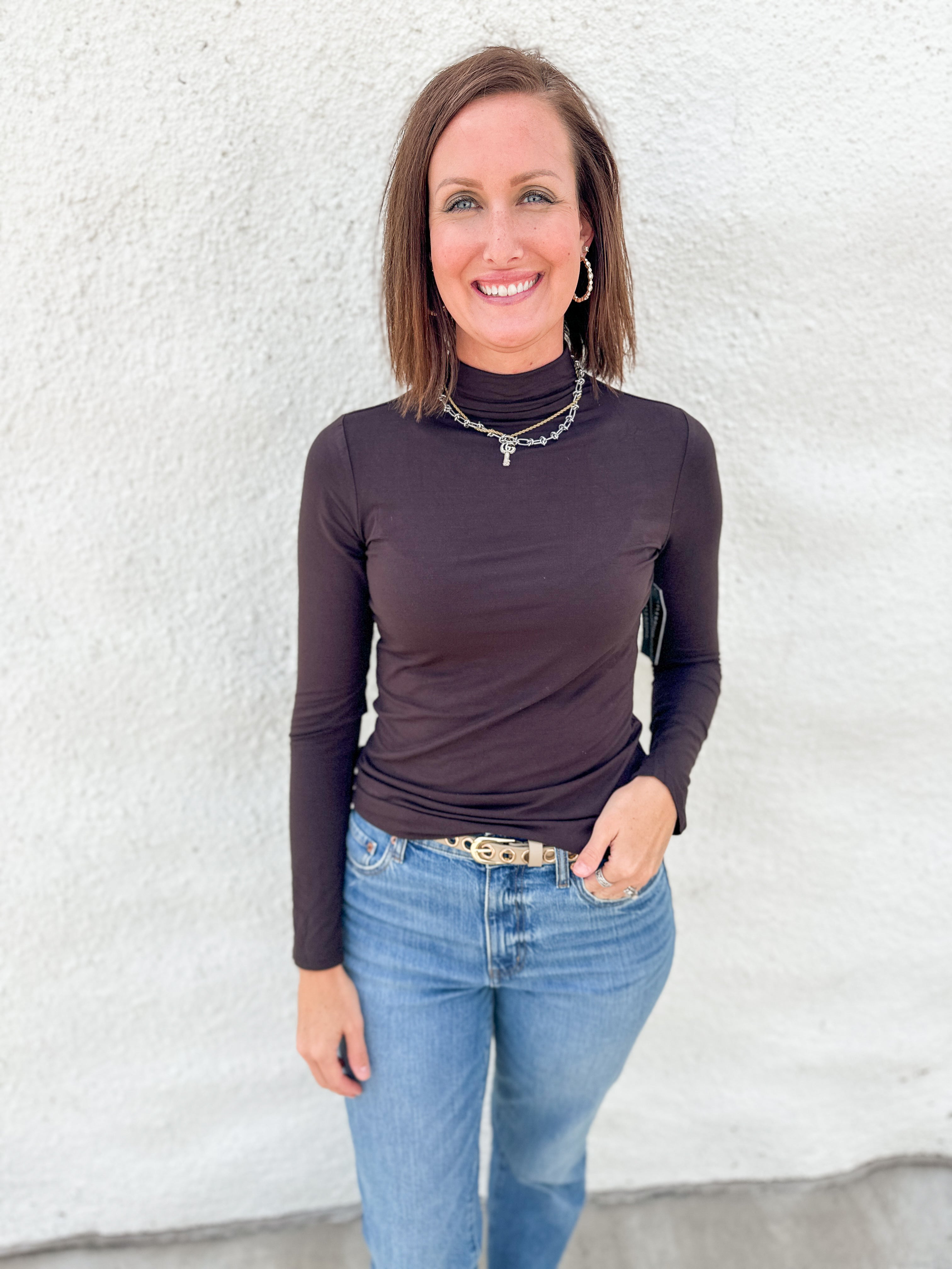 A woman wearing a dark brown long-sleeve mock neck turtleneck top and blue jeans, standing against a white textured wall.