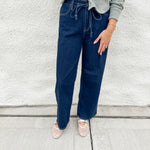 A person wearing dark blue wide-leg drawstring jeans, a light green cardigan, and cream mesh mary jane flats, standing against a white textured wall.