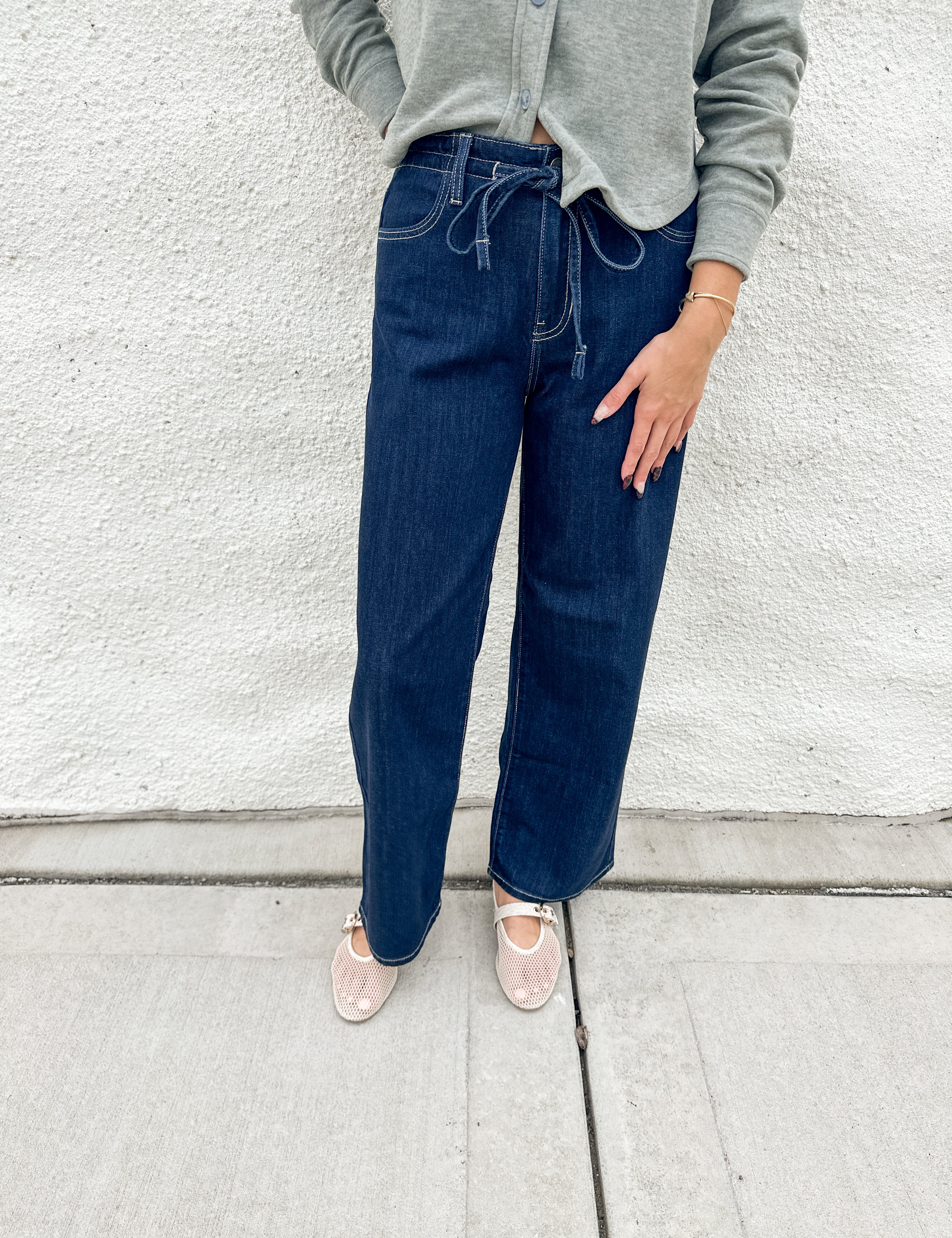 A person wearing dark blue wide-leg drawstring jeans, a light green cardigan, and cream mesh mary jane flats, standing against a white textured wall.