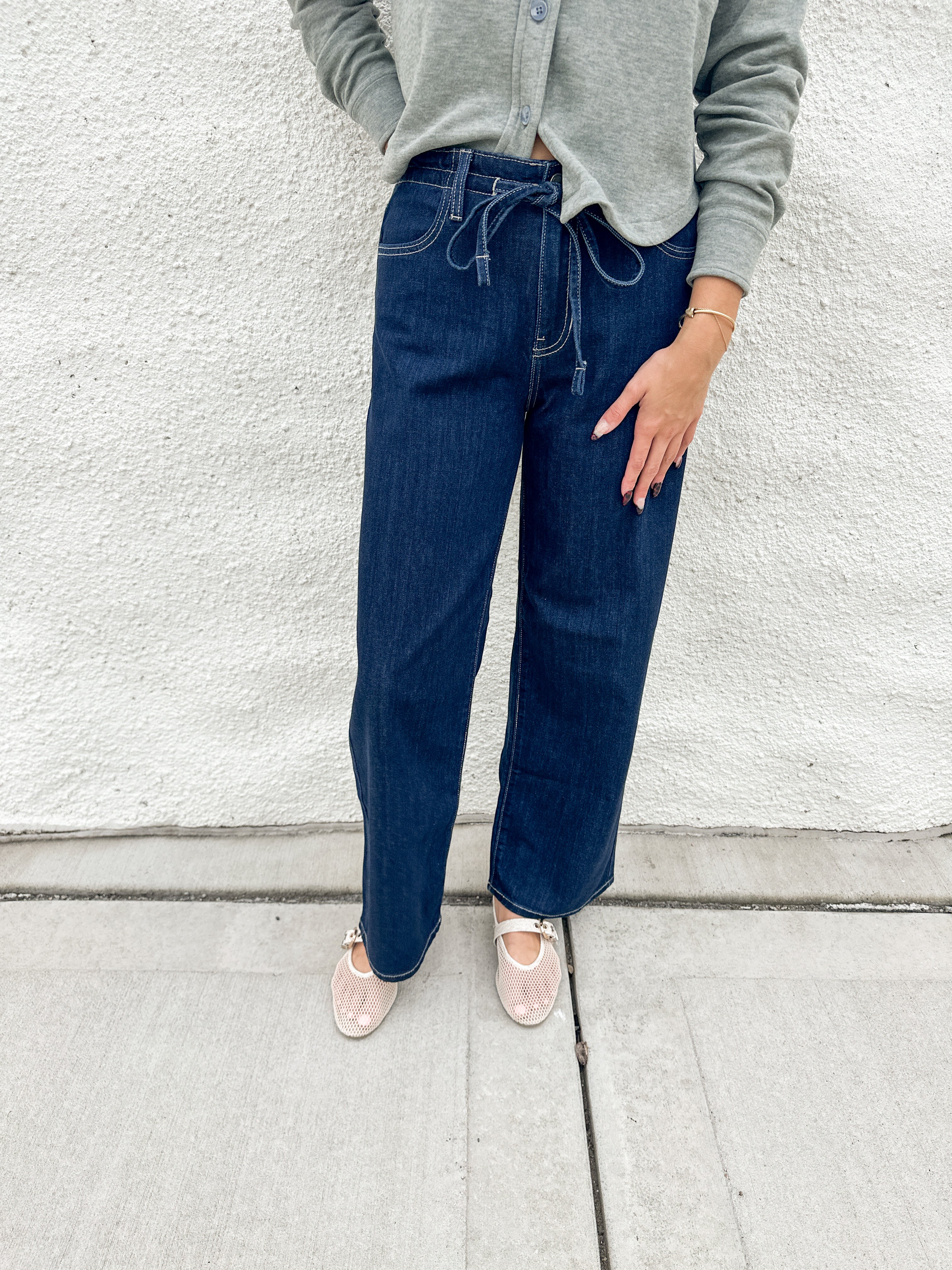 A person wearing dark blue wide-leg drawstring jeans, a light green cardigan, and cream mesh mary jane flats, standing against a white textured wall.