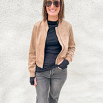 A woman wearing a tan suede bomber jacket over a black turtleneck, gray jeans, and sunglasses, standing against a white textured wall.