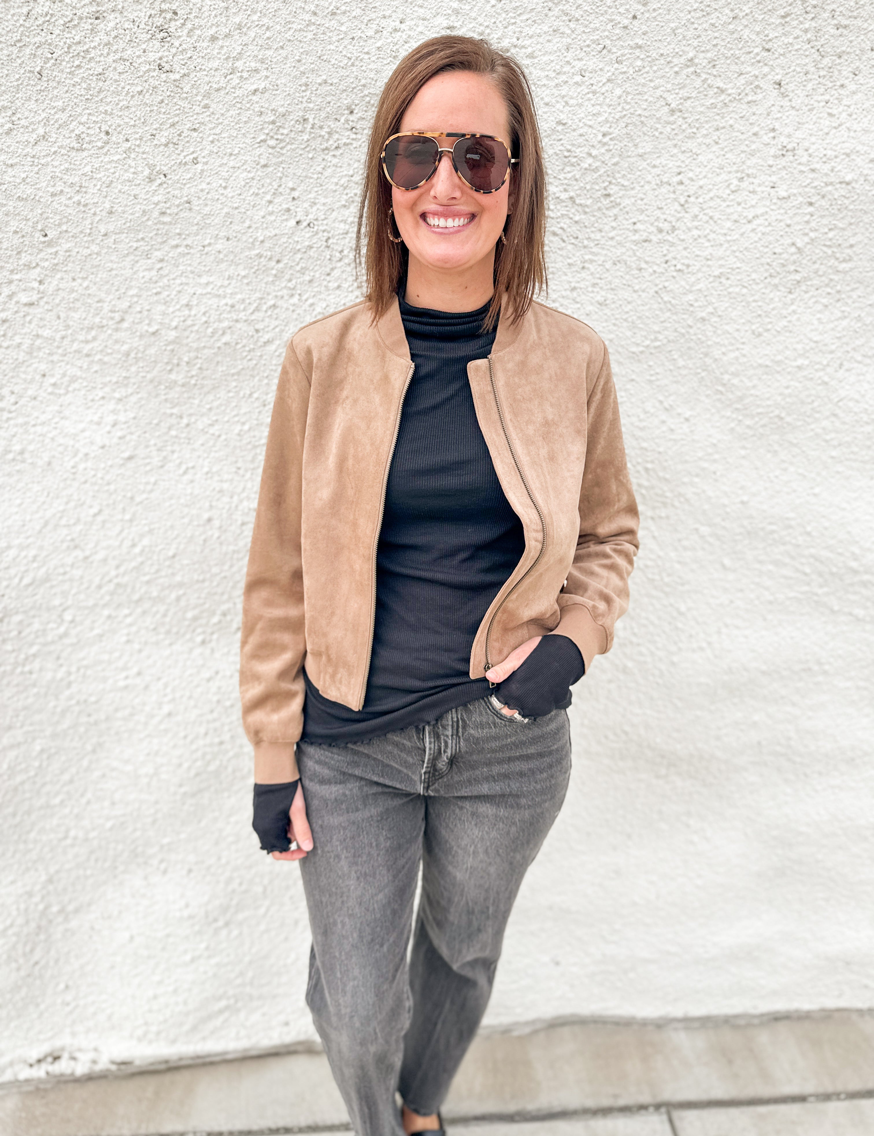 A woman wearing a tan suede bomber jacket over a black turtleneck, gray jeans, and sunglasses, standing against a white textured wall.