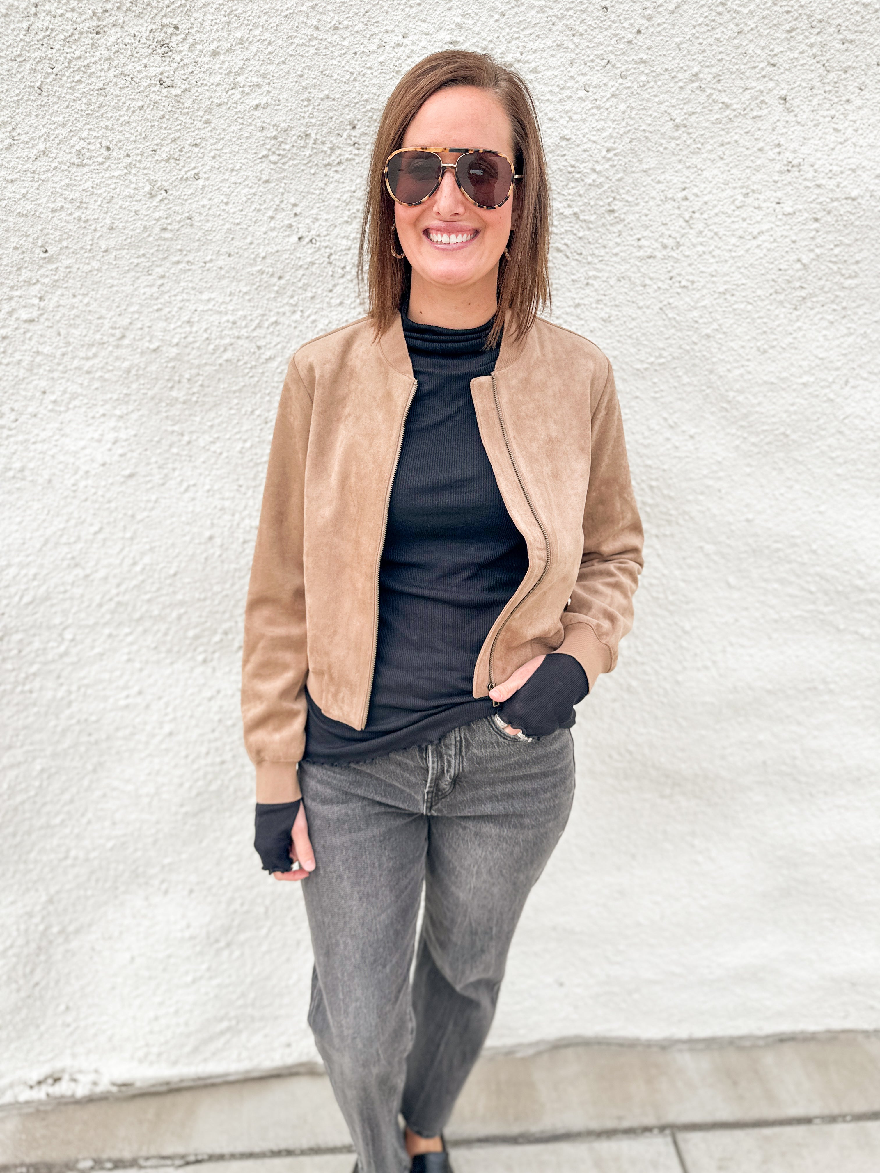 A woman wearing a tan suede bomber jacket over a black turtleneck, gray jeans, and sunglasses, standing against a white textured wall.