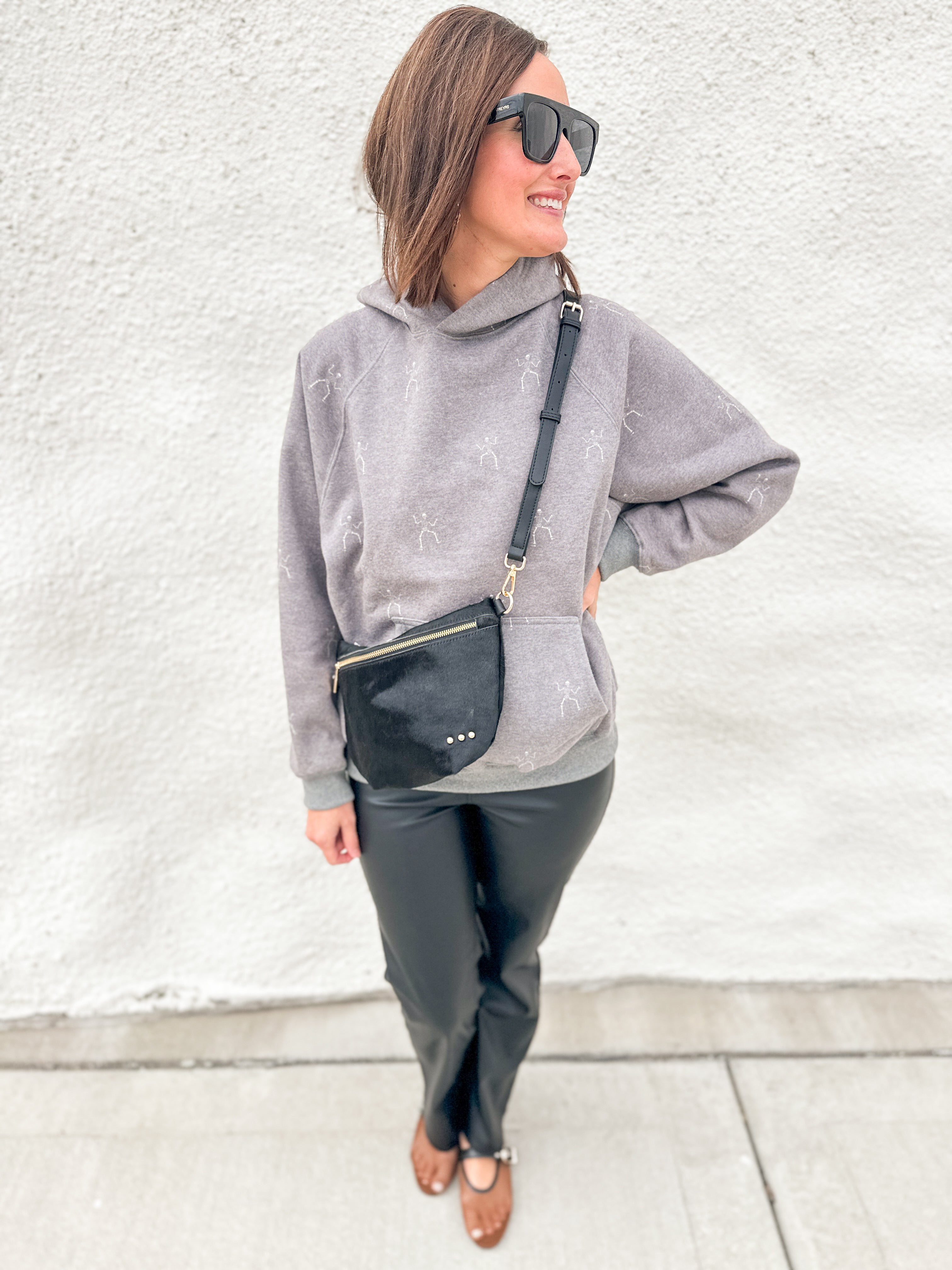 A woman wearing a light gray hooded sweatshirt with a subtle tonal pattern, black faux-leather pants, and brown flat shoes, carrying a black crossbody bag with gold hardware against a white stucco wall.