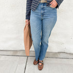 A person wearing blue straight-leg jeans, a navy and black plaid blazer over a white top, brown mesh Mary Jane flats, and holding a tan leather bag, standing against a white stucco wall.