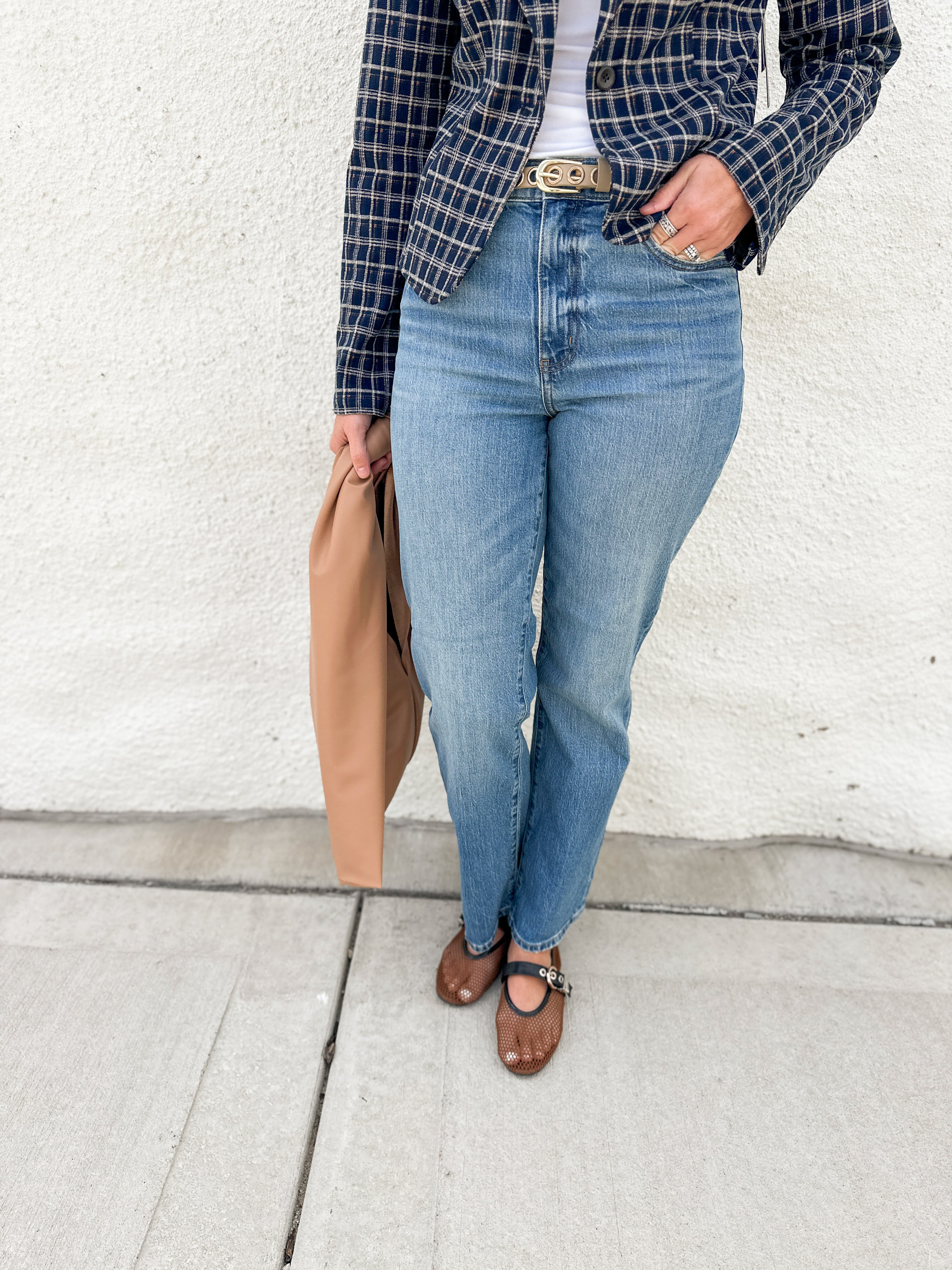 A person wearing blue straight-leg jeans, a navy and black plaid blazer over a white top, brown mesh Mary Jane flats, and holding a tan leather bag, standing against a white stucco wall.