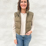 A woman wearing an olive green cropped quilted vest layered over a cream sweater wearing blue jeans in front of a white wall