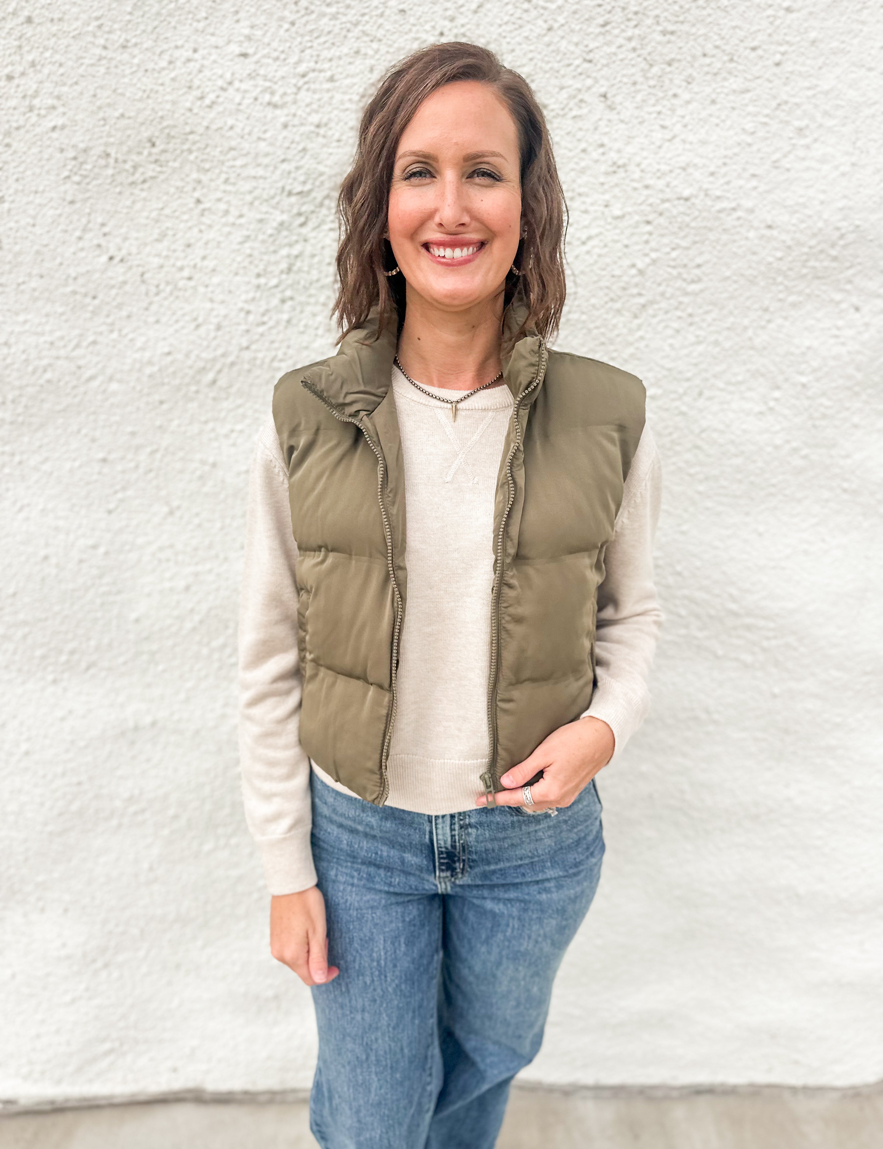 A woman wearing an olive green cropped quilted vest layered over a cream sweater wearing blue jeans in front of a white wall