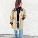 A woman wearing a olive barn jacket trench coat with dark brown corduroy collar and cuffs, styled with a brown turtleneck, blue jeans, and brown mesh mary jane flats, standing against a white textured wall.