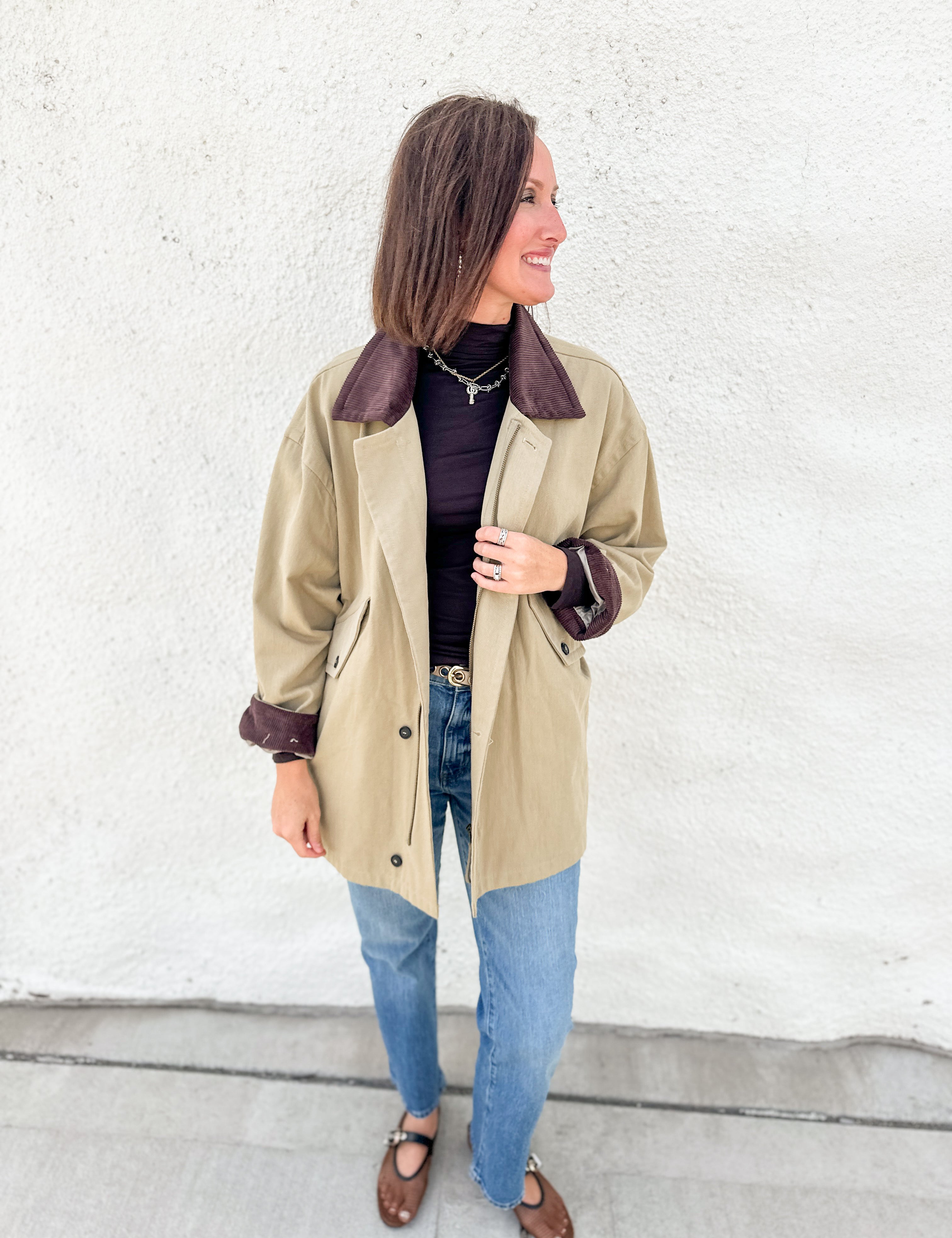 A woman wearing a olive barn jacket trench coat with dark brown corduroy collar and cuffs, styled with a brown turtleneck, blue jeans, and brown mesh mary jane flats, standing against a white textured wall.