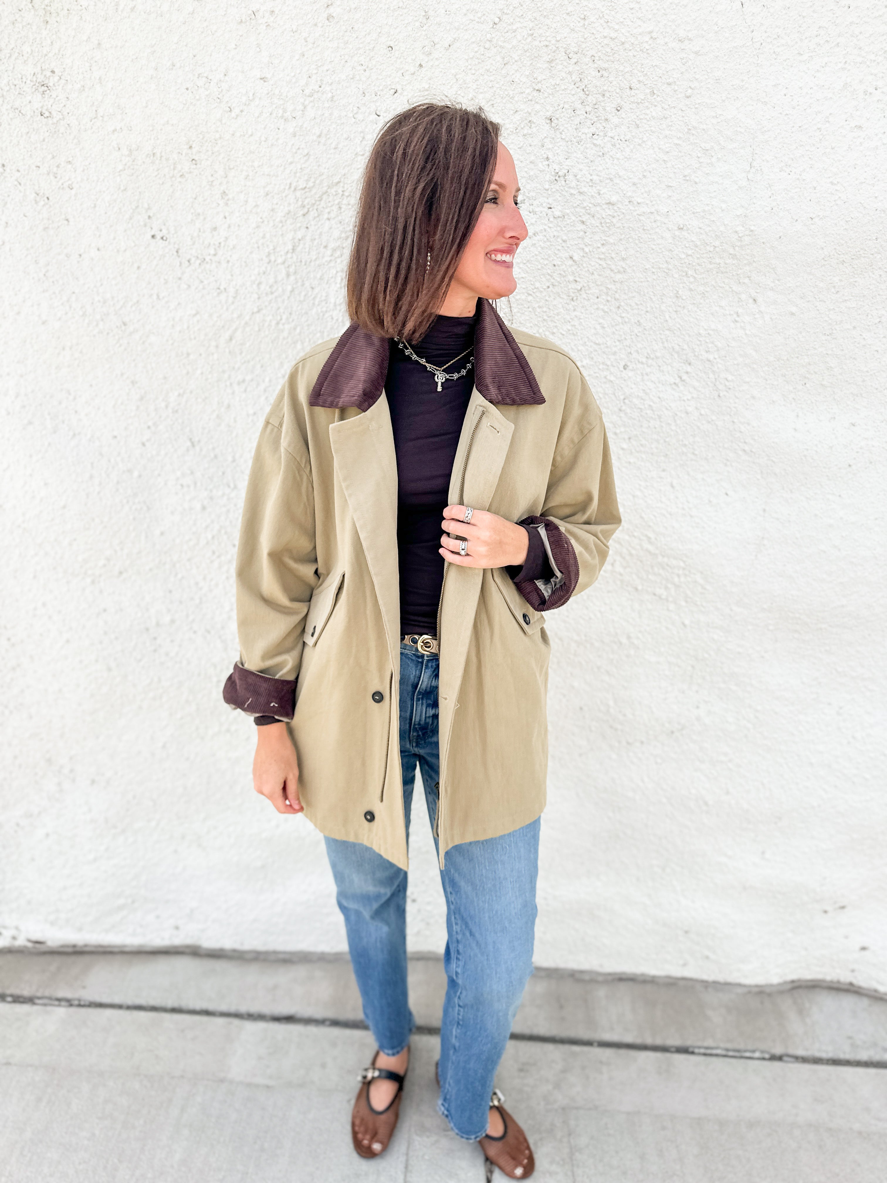 A woman wearing a olive barn jacket trench coat with dark brown corduroy collar and cuffs, styled with a brown turtleneck, blue jeans, and brown mesh mary jane flats, standing against a white textured wall.