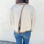 A person viewed from behind wearing a cream-colored knit sweater with a brown trim detail running down the center bac open back detail, paired with blue jeans, standing against a white stucco wall.
