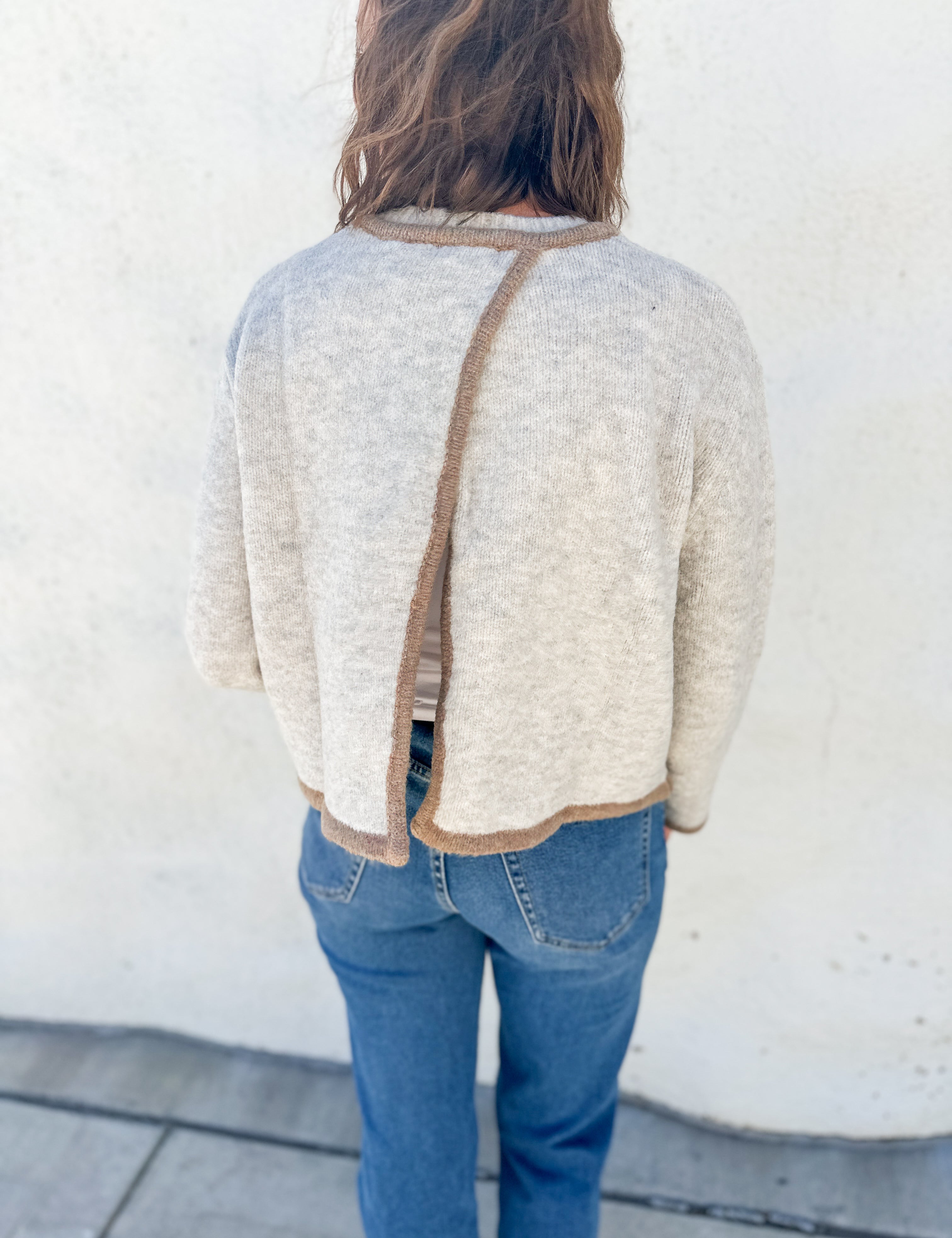 A person viewed from behind wearing a cream-colored knit sweater with a brown trim detail running down the center bac open back detail, paired with blue jeans, standing against a white stucco wall.