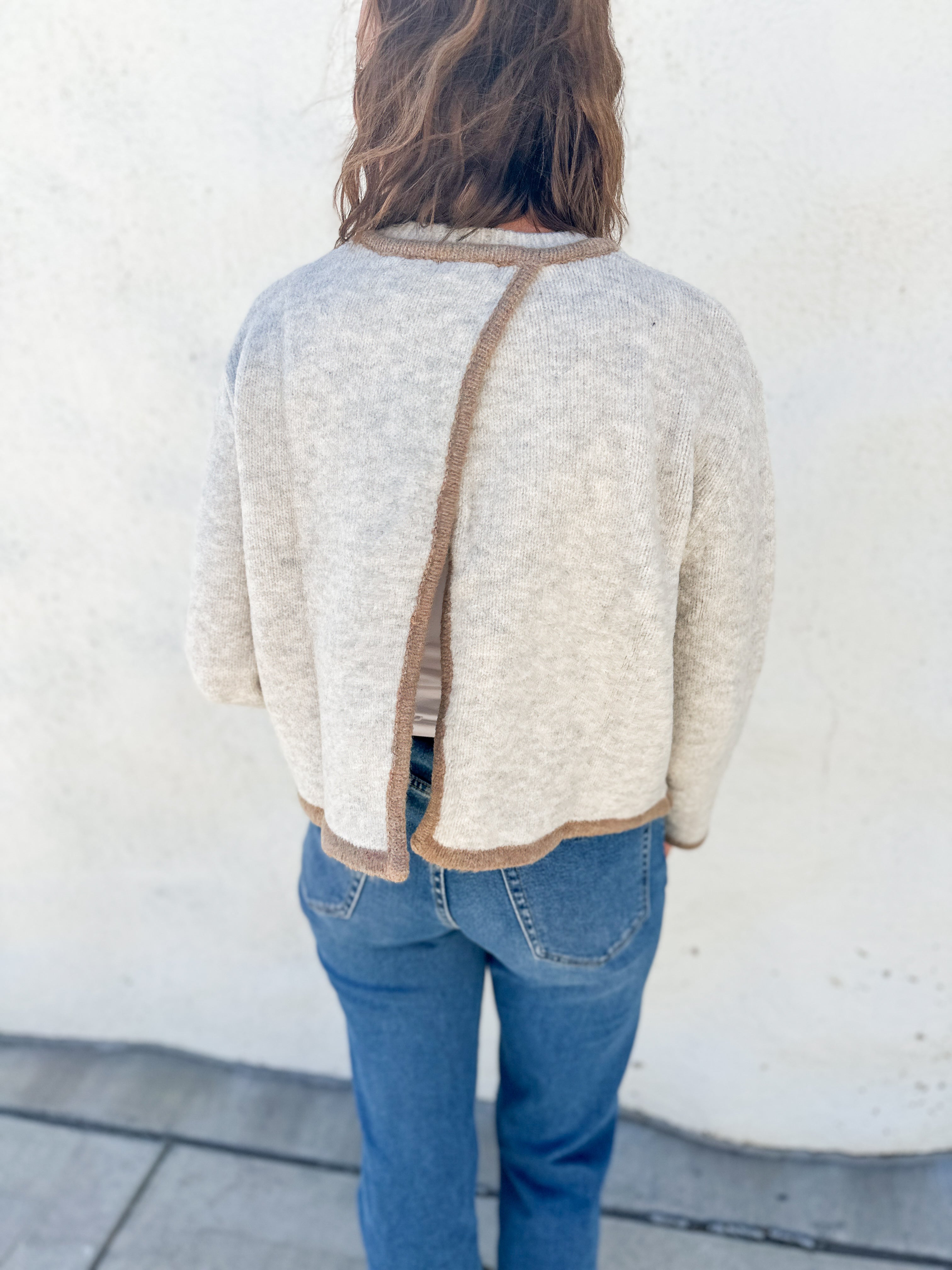 A person viewed from behind wearing a cream-colored knit sweater with a brown trim detail running down the center bac open back detail, paired with blue jeans, standing against a white stucco wall.