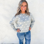 A woman with gray hair stands against a white stucco wall wearing a light gray long-sleeve knit sweater with white polka dots and blue jeans.